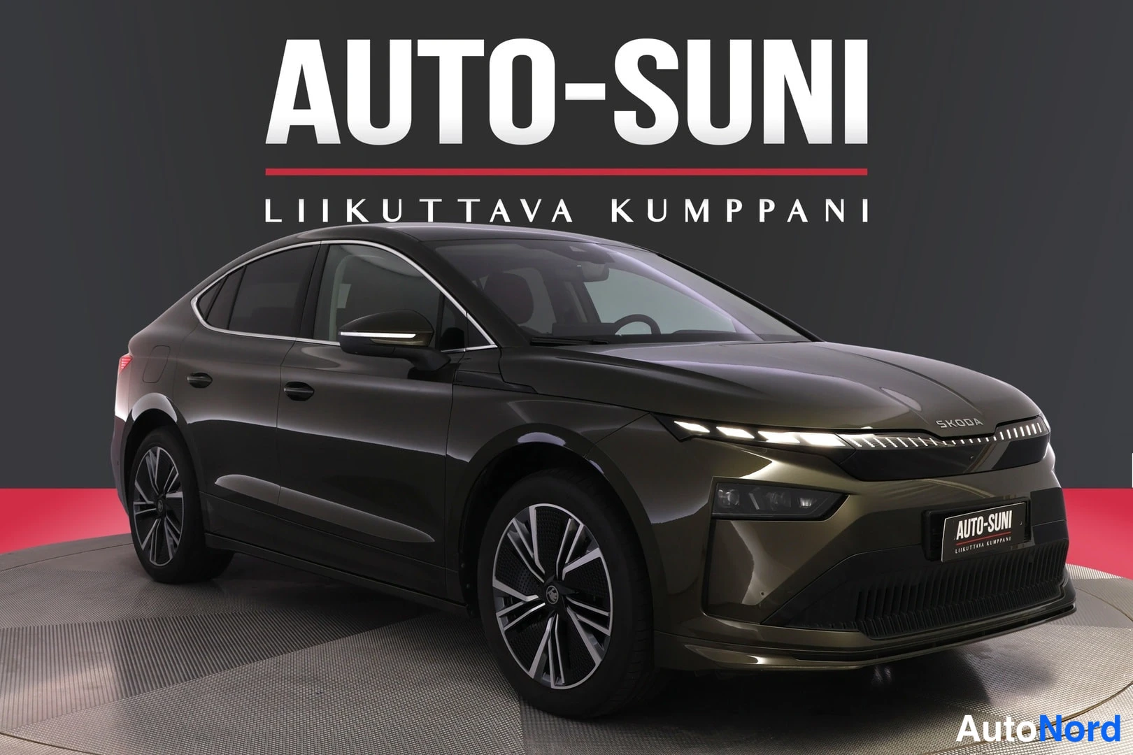 Etuviisto, nokka oikealle: Skoda Enyaq Coupé tummanruskeassa värissä, etukulmakuva studiotaalustossa Auto-Suni-logon alla.