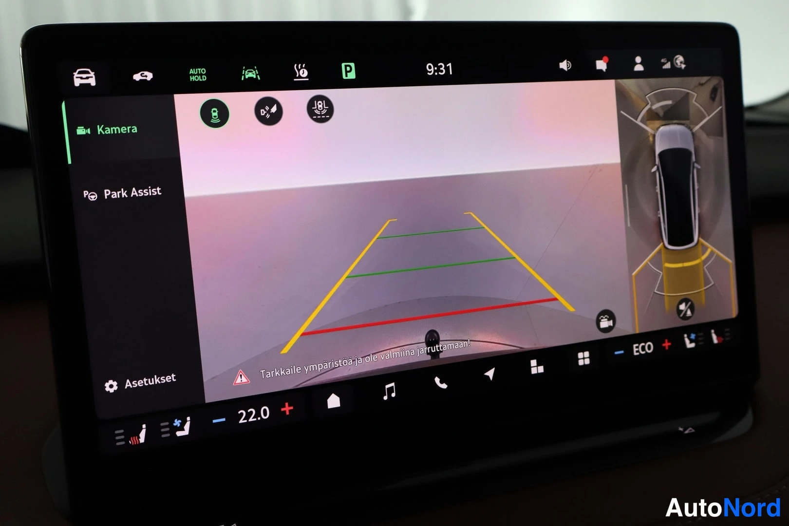 Yksityiskohta: Infotainment-näytön Kamera-näkymä pysäköintikameralla ja Park Assist-toiminnolla; opasteet tarkkaile ympäristöä ennen jarruttamista.