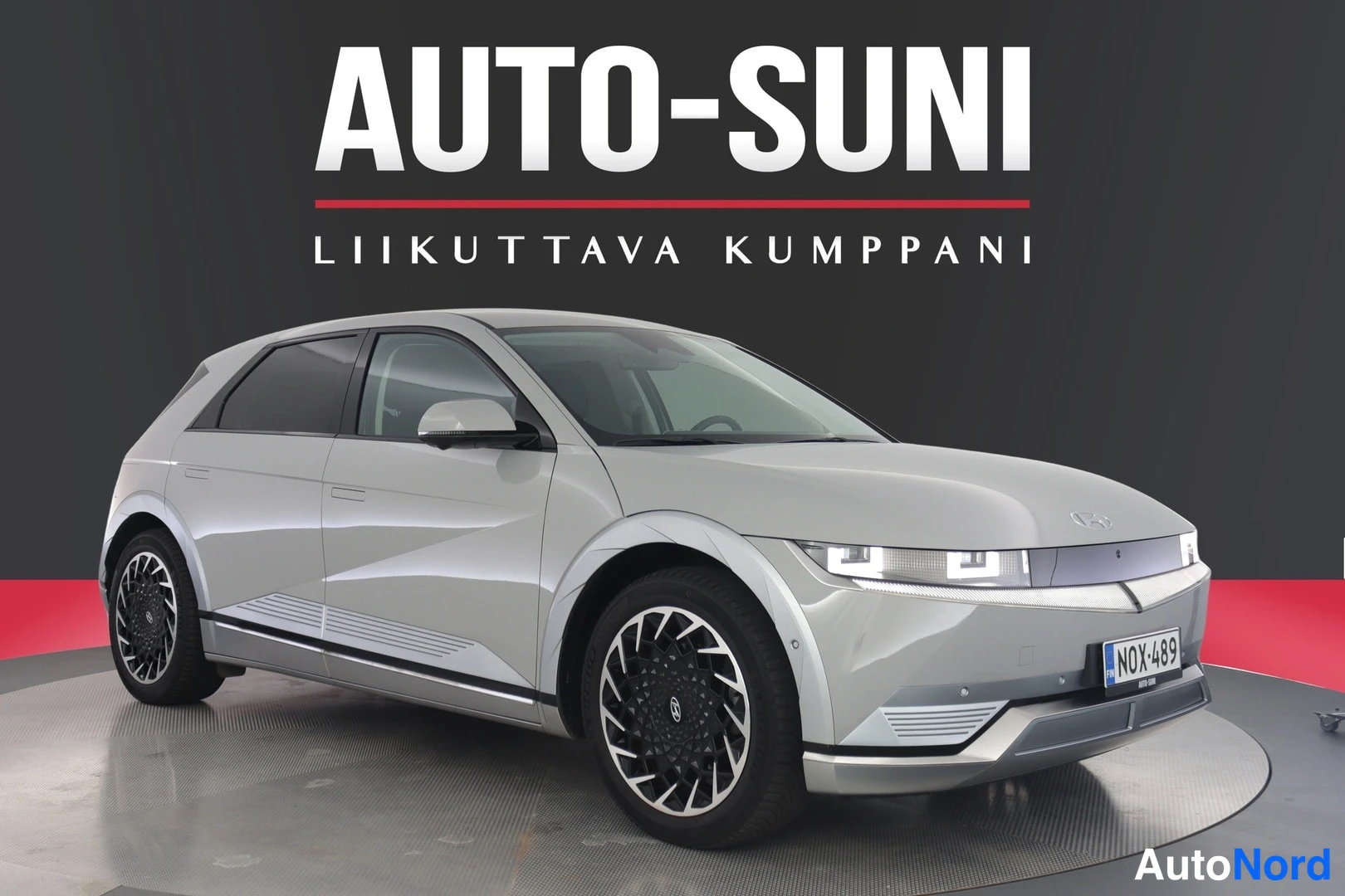 Etuviisto, nokka vasemmalle: Front three-quarter view of the silver car with black multi-spoke alloy wheels visible. LED headlights are on.