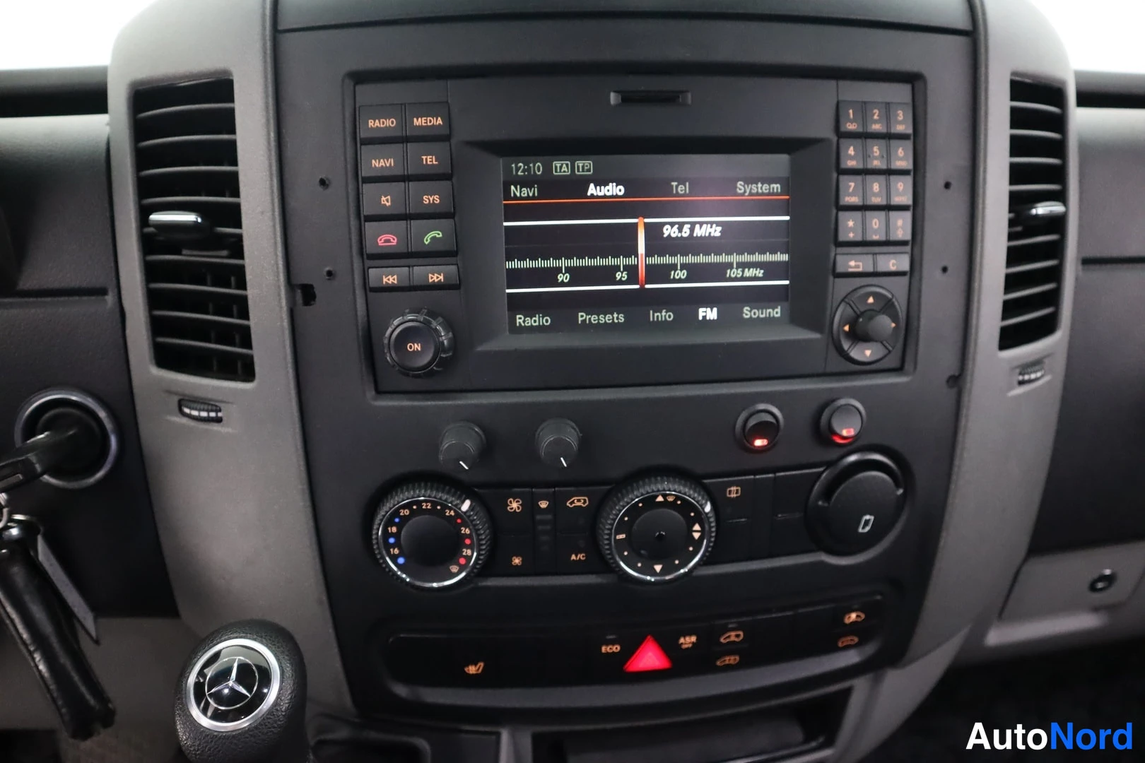 Yksityiskohta: Mercedes-merkkisen keskikonsolin radio- ja navigointiyksikkö näytöllä 96.5 MHz, ympärilläl näppäimistö ja painikkeet, alapuolella lämpötilan säätimet ja ECO/ASR