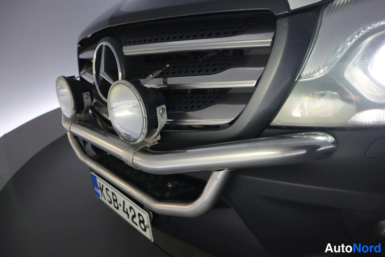 Edestä: Mercedes-Benz Sprinterin etuosan lähikuva: merkkitunnus keulalla, kromattu suojaraudanne ja kaksi lisävaloa, rekisteritunnus.