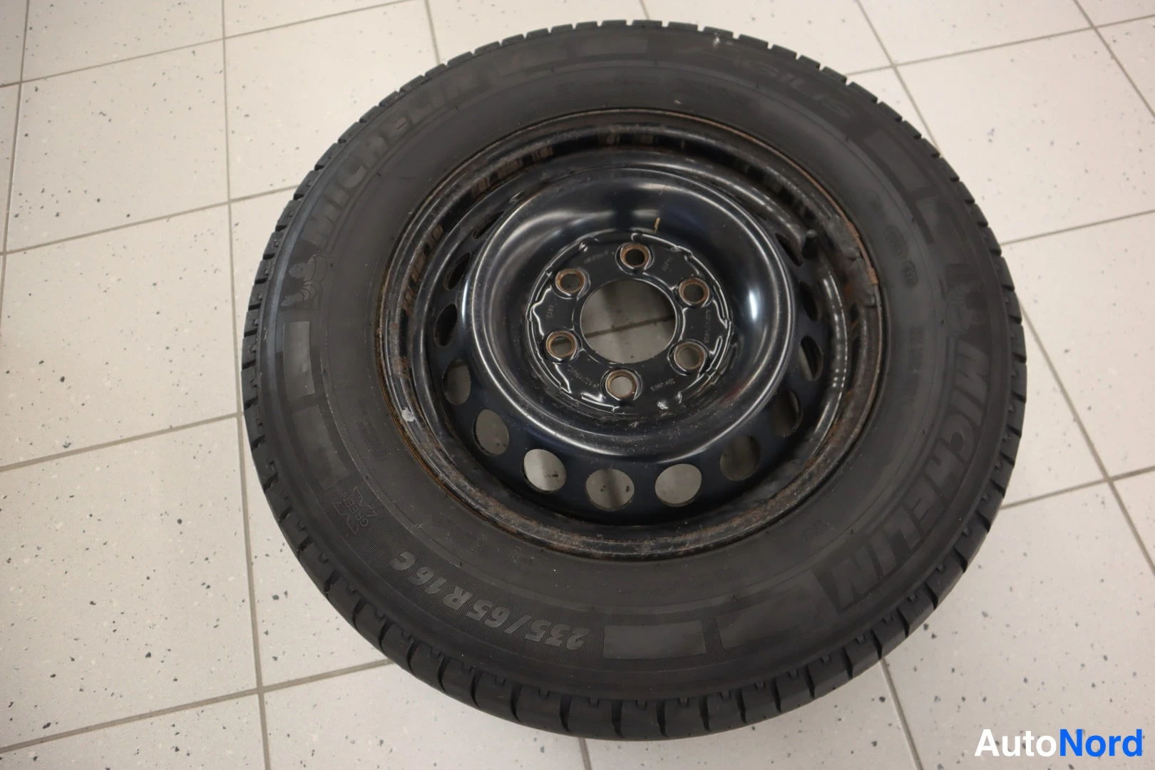 Yksityiskohta: Varapyörä lattialla: musta teräsvanne ja Michelin-merkkinen rengas, kokomerkintä 235/65 R16 C nähtävissä.
