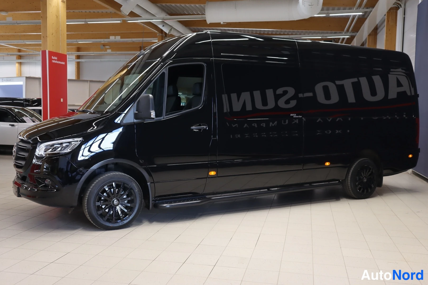 Sivulta: Musta Mercedes-Benz Sprinter -pakettiauto etuviistosta vasempaan kulmassa sisätiloissa, kyljessä myyntiautoketjun heijastus.