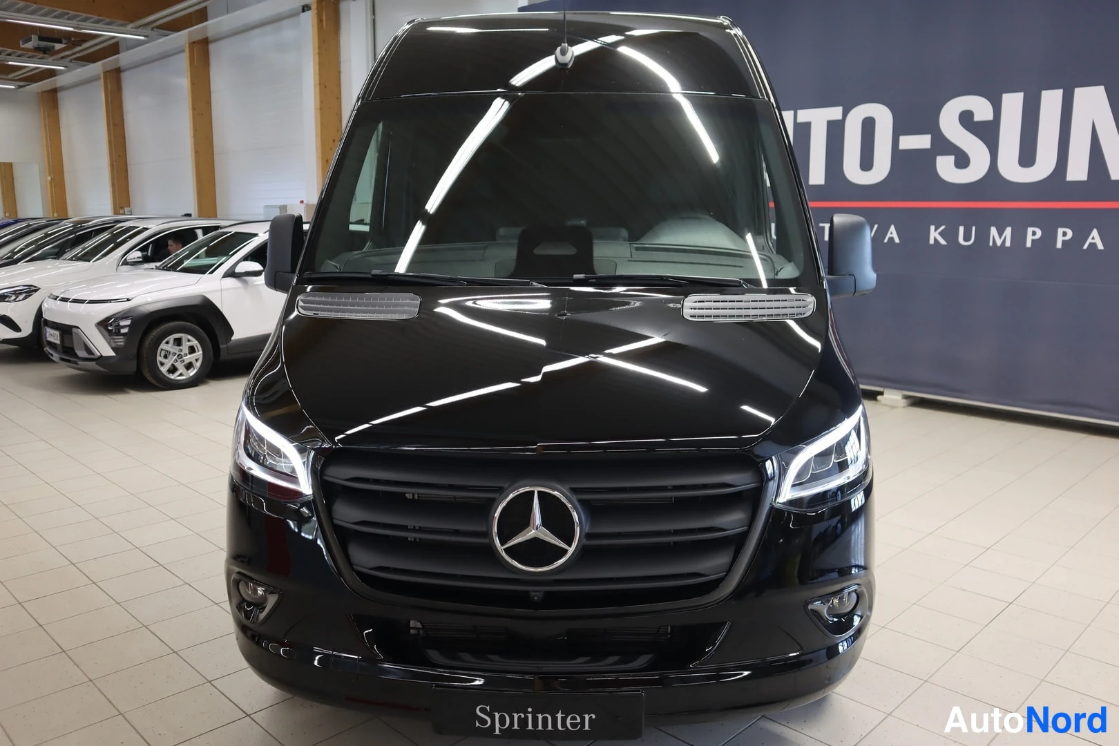 Edestä: Musta Mercedes-Benz Sprinter -pakettiauto suoraan edestä, keulalla Mercedes-tähti ja Sprinter-teksti.