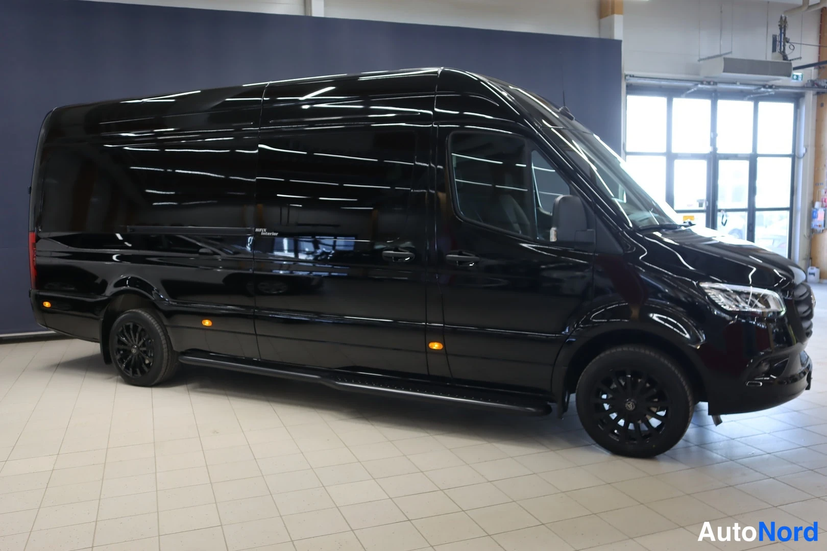 Etuviisto, nokka oikealle: Musta pitkä Mercedes-Benz Sprinter -pakettiauto etuviistosta oikealta, mustat kevytmetallivanteet ja sivukaiteet.