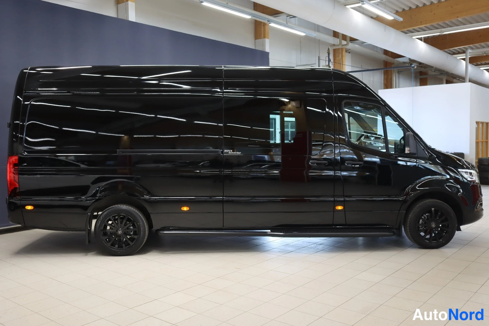 Sivulta: Mustan Mercedes-Benz Sprinter -pakettiauton sivuprofiili oikealta sisätiloissa, kromattu.lista ja mustat vanteet näkyvät.