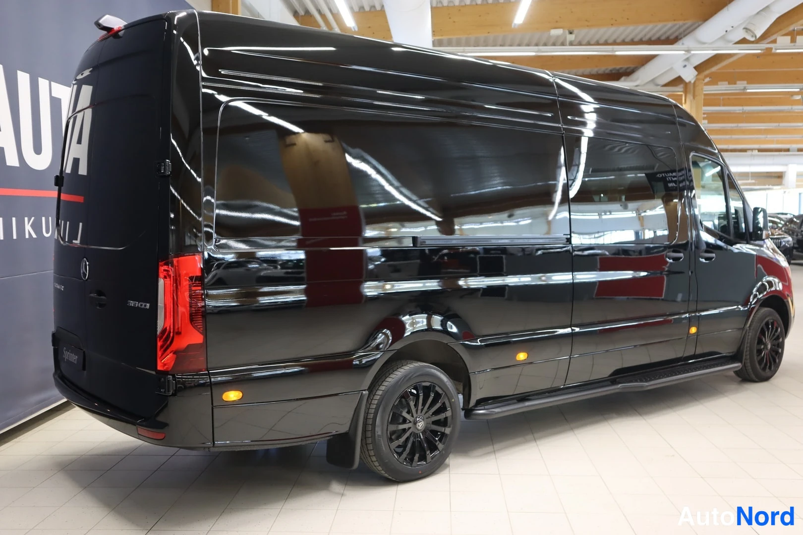 Takaviisto: Musta Mercedes-Benz Sprinter -pakettiauto takaviistosta, takavalot ja autoc:symboli näkyvät.