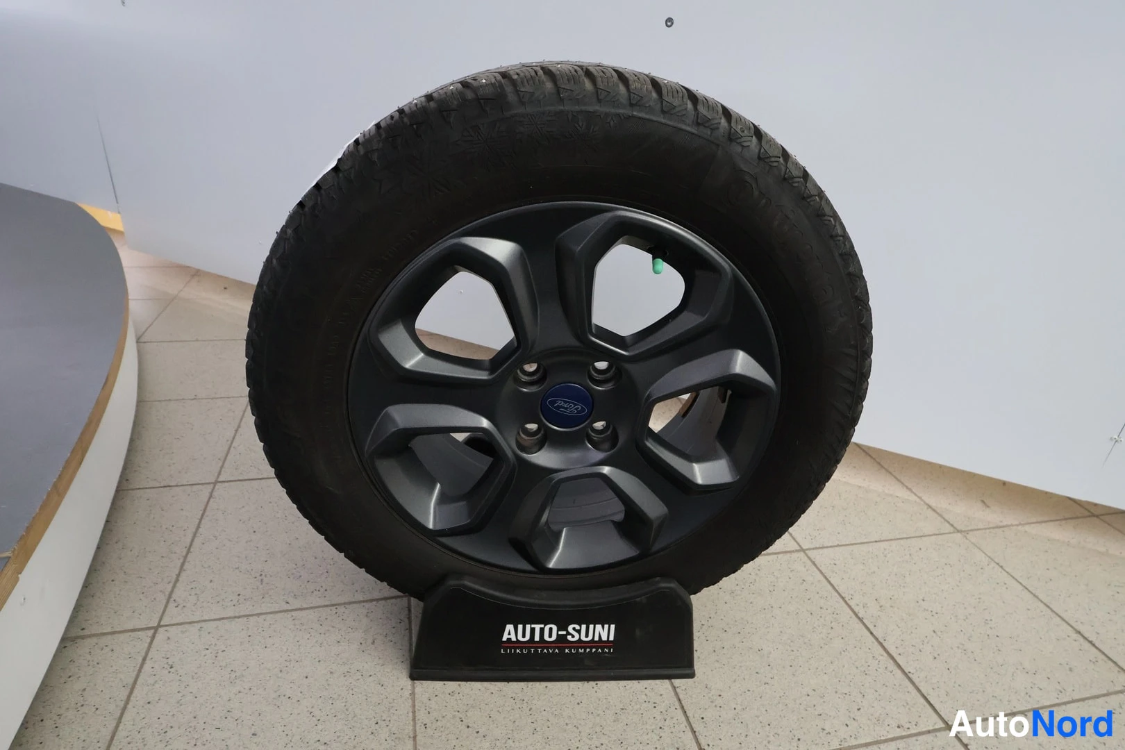Yksityiskohta: Käytetty rengas tummalla Ford-levyvanteella esitteellä Auto-Suni-telineessä sisätiloissa.