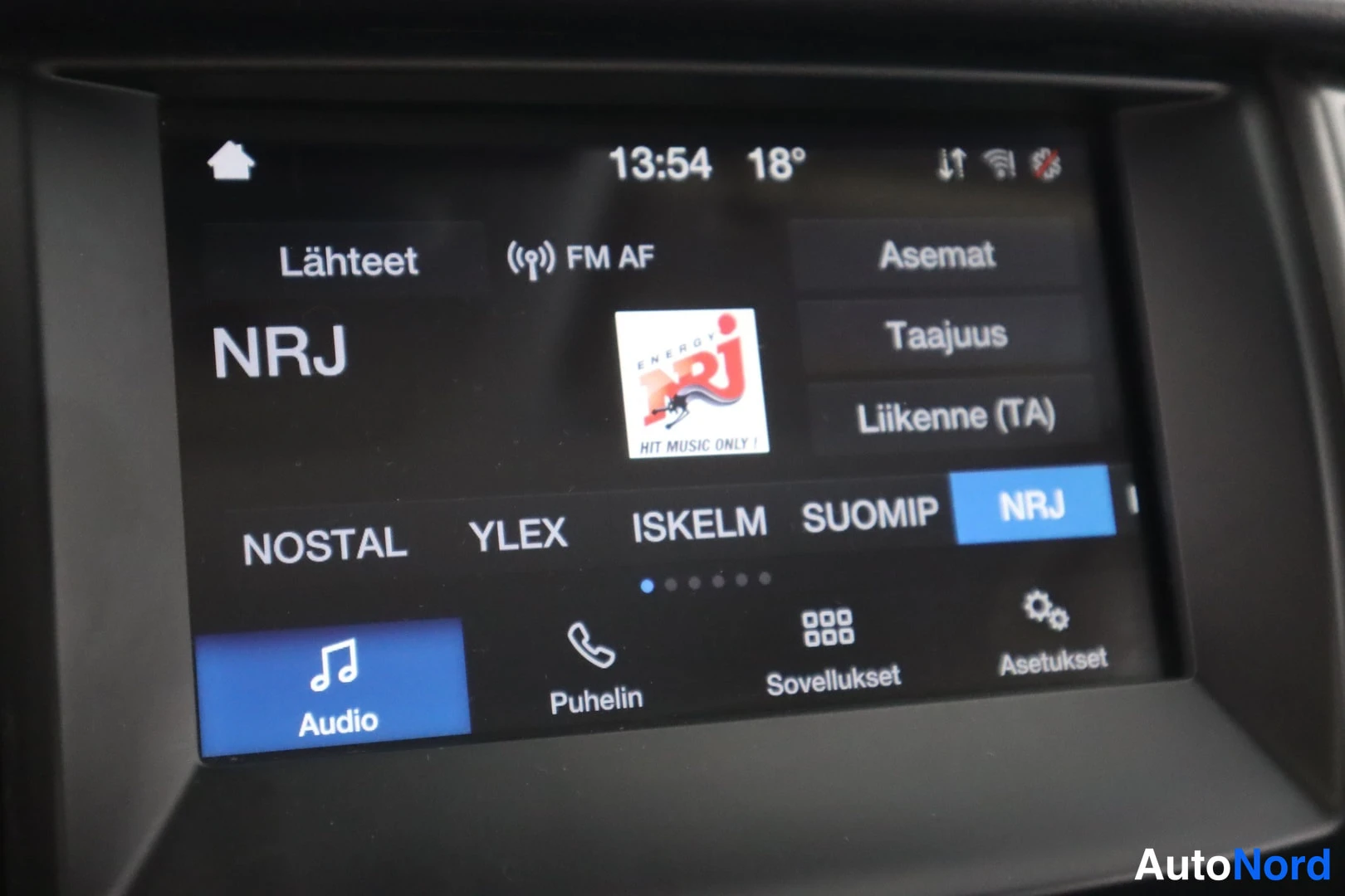 Yksityiskohta: Kojelaudan kosketusnäyttö lähikuvana, jossa radiovalikko: valittuna kanava NRJ, kellonaika 13:54 ja lämpötila 18 astetta. Ruudulla valikot Lähteet, Asemat, Taaj