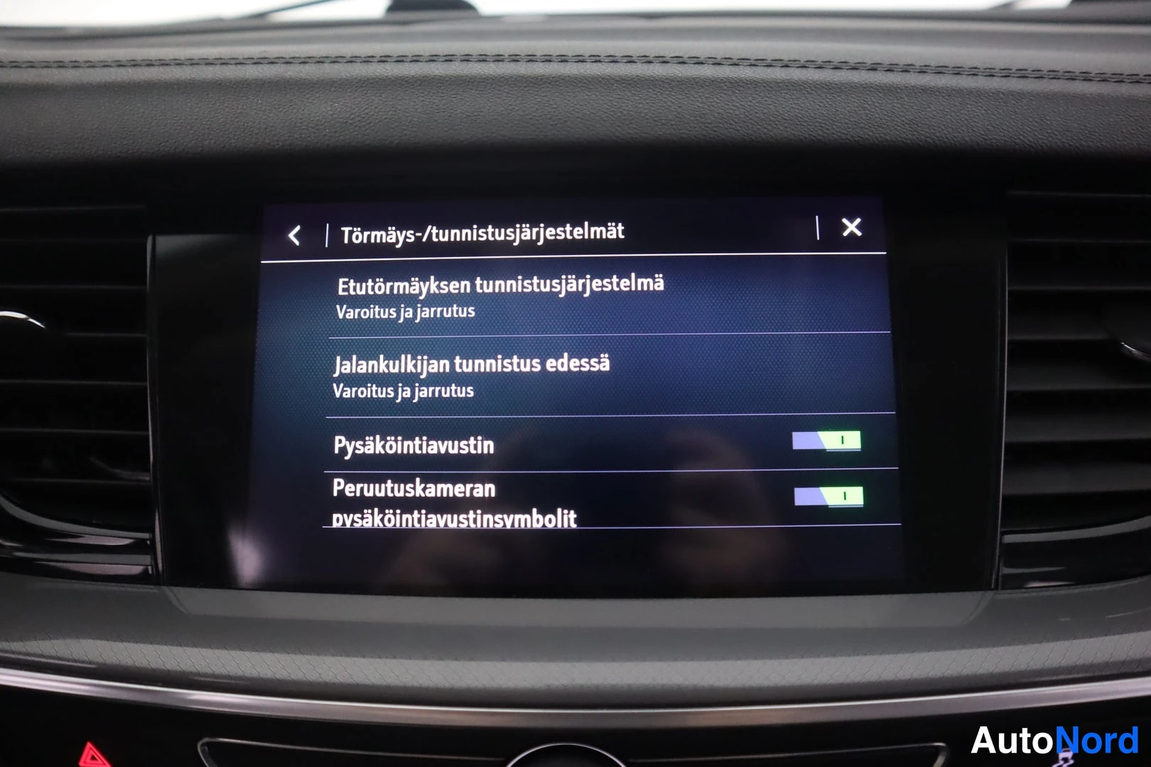Yksityiskohta: Auton kojelaudan infotainment-näyttö, jossa Törmäys-/tunnistusjärjestelmät-valikko mm. etutörmäyksen tunnistusjärjestelmä ja peruutuskameran pysäköintiaustinsym