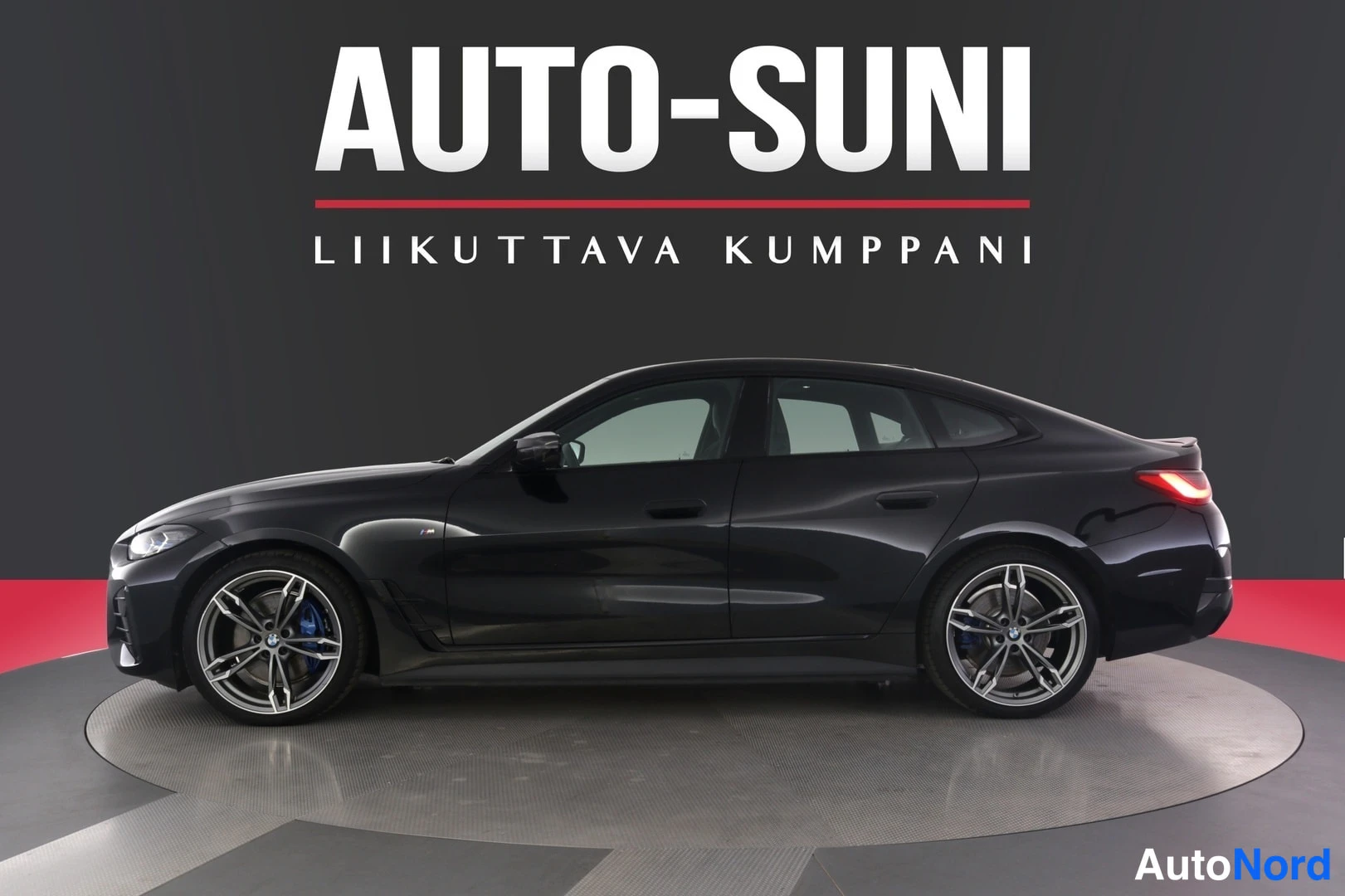 Etuviisto, nokka vasemmalle: Autosuni-myynnissä oleva musta BMW i4 neliovinen coupe nähtynä vasemmasta etukulmasta tummassa studiossa punaisella lattialla