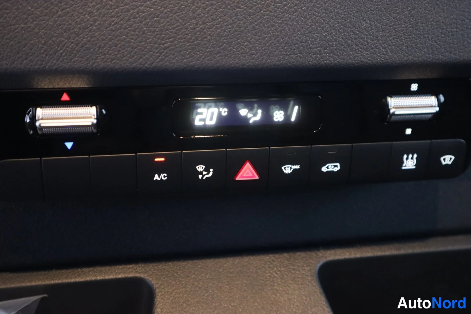 Yksityiskohta: Kuva kojelaudan ilmastointipaneelista, jossa näkyy digitaalinen lämpötila-asetus 20°C, kaksi ilmastointisäädintä ja painikkeet kuten A/C, plyysi-ikoni, punainen