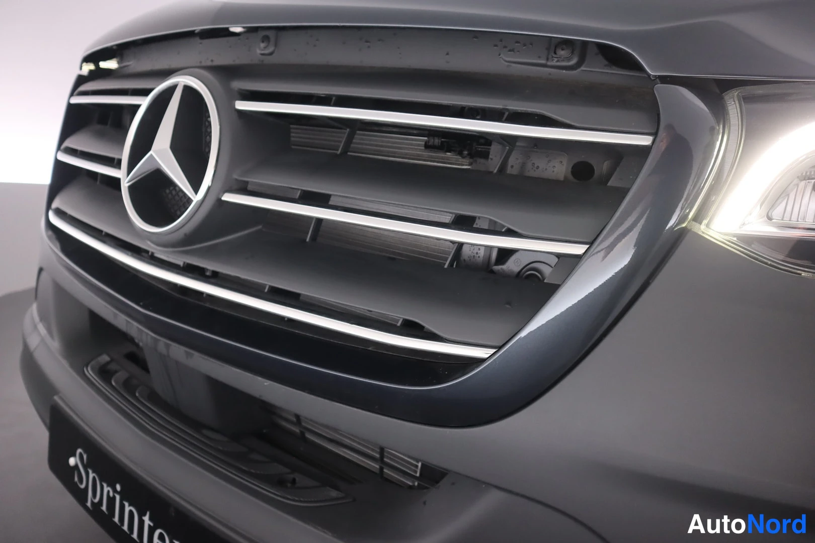 Edestä: Lähikuva Mercedes-pakettiauton etusäleiköstä ja Mercedes-tähtimerkistä; kiiltävä tumma kori ja osa etuvalaisimesta näkyvissä.