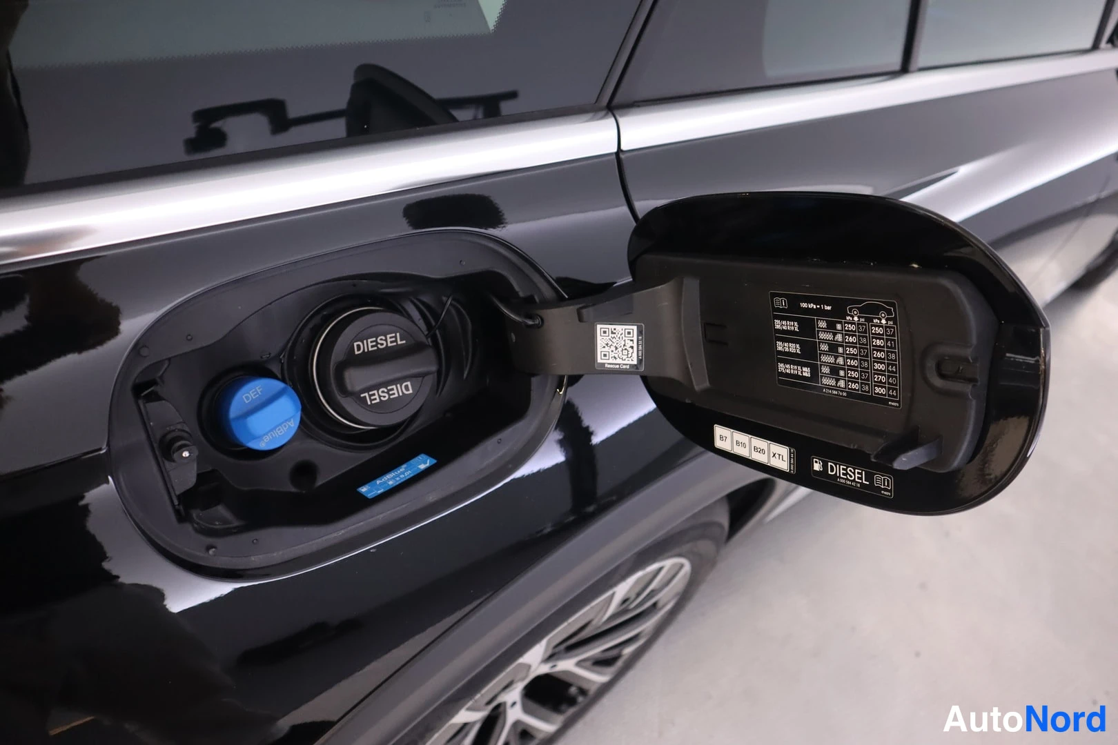 Yksityiskohta: Mustan auton avattu tankkiluukku: musta DIESEL-tankatulppa ja sininen AdBlue-täyttöaukko, luukun sisäpuolella taulukkolappu ja DIESEL-merkintä.