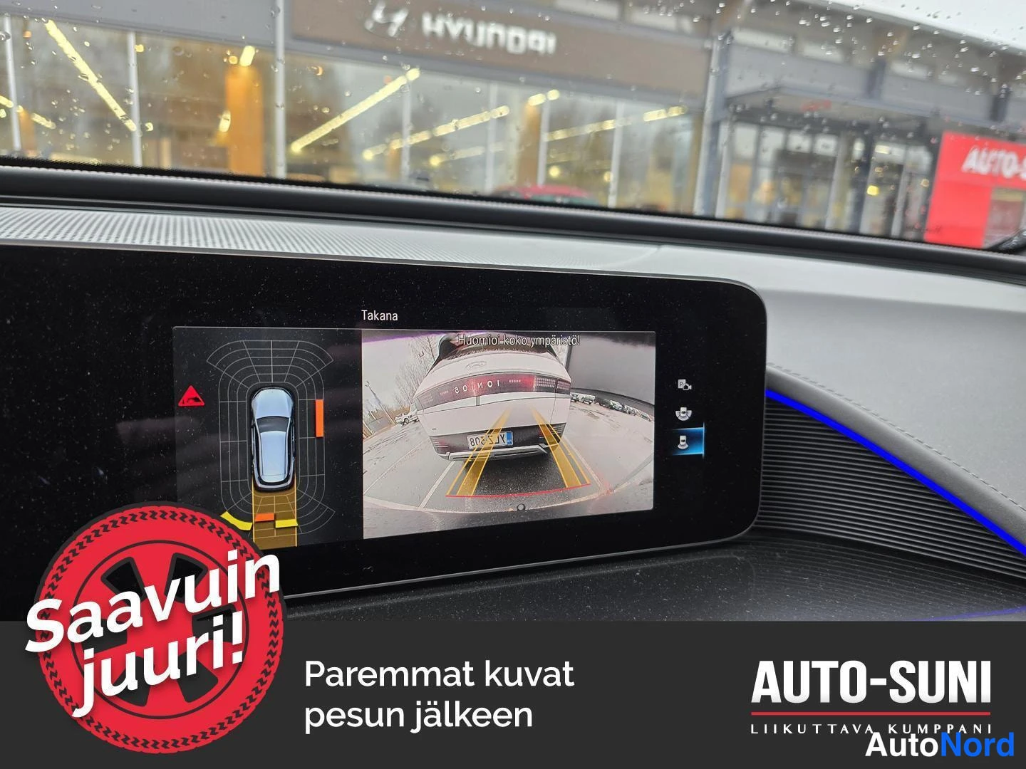 Ohjaamo: Keskuspaneelin näyttö näyttää takapainokuvaan liittyvän kuvaohjelman ja taustakuvia. Takavaloissa näkyy toisen auton kuva. Kuvan alareunassa on logo 'Saavuin ju
