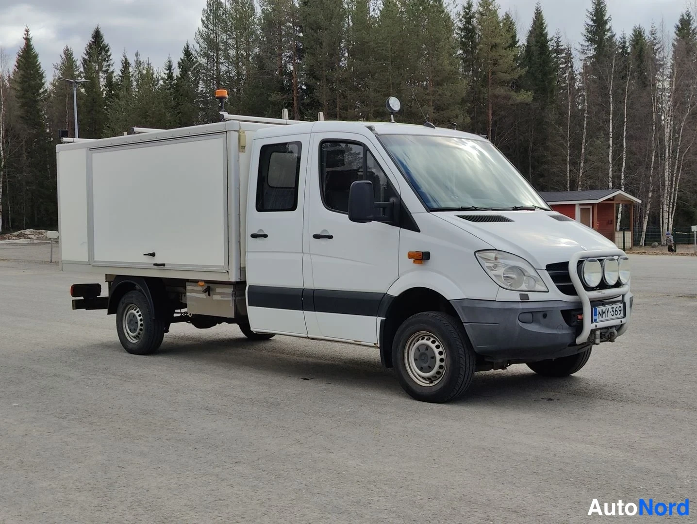 Etuviisto, nokka oikealle: Valkoinen Mercedes-Benz Sprinter -työauto neliovinen kabiinin ja takana olevan erilliskorin ulkonäkö. Etuvaloissa on lisävalot