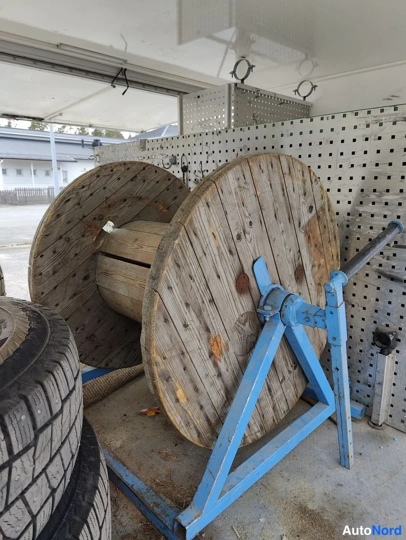 Yksityiskohta: Detail shot inside the cargo area focusing on equipment. Large wooden industrial cable reels rest on blue painted metal stands. Partial view of tires with winte