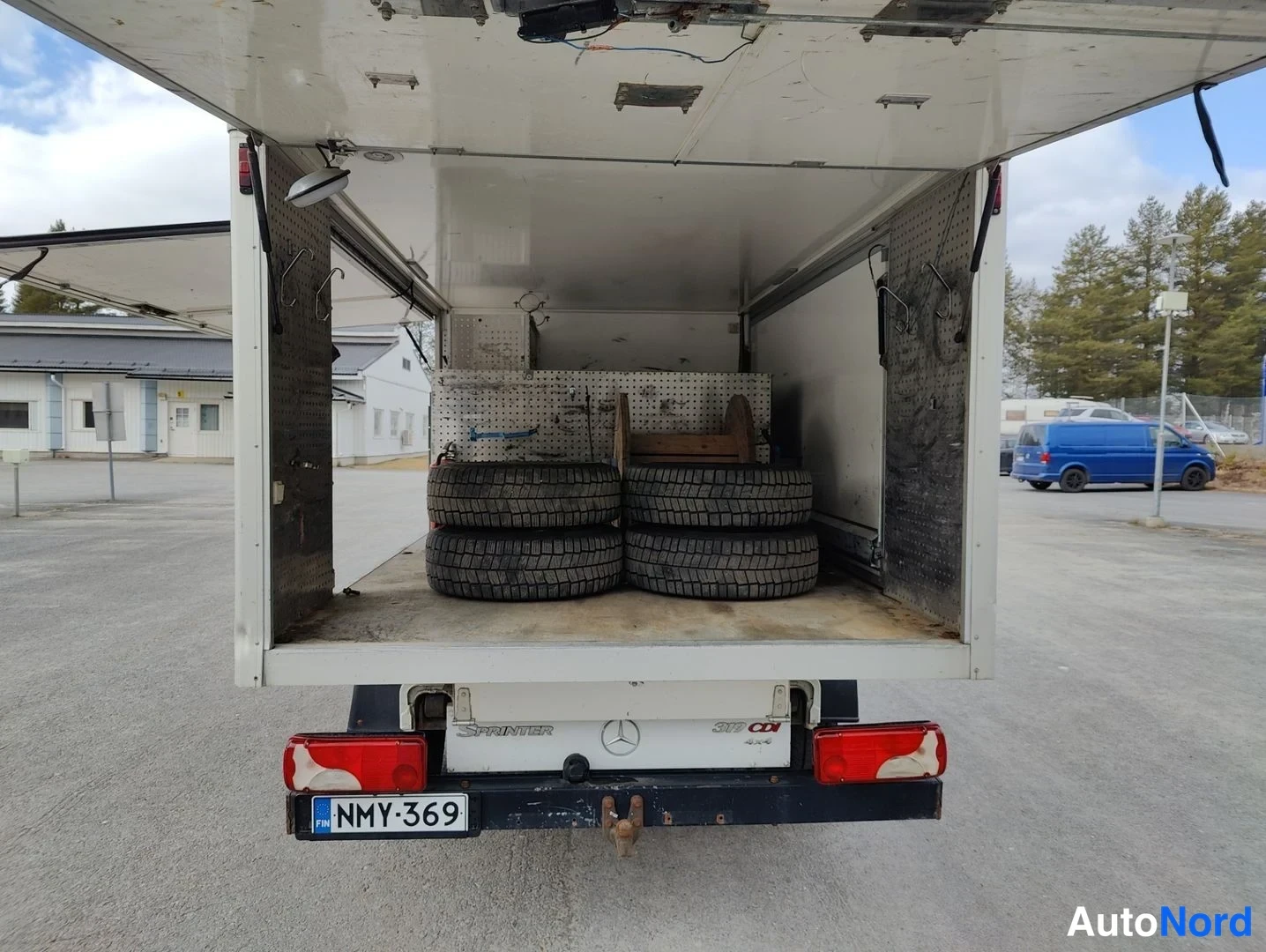 Takaviisto: Rear three-quarter view of the truck showing the open lift-gate cargo bed filled with tires and a metal toolbox. The Mercedes-Benz Sprinter tailgate is visible