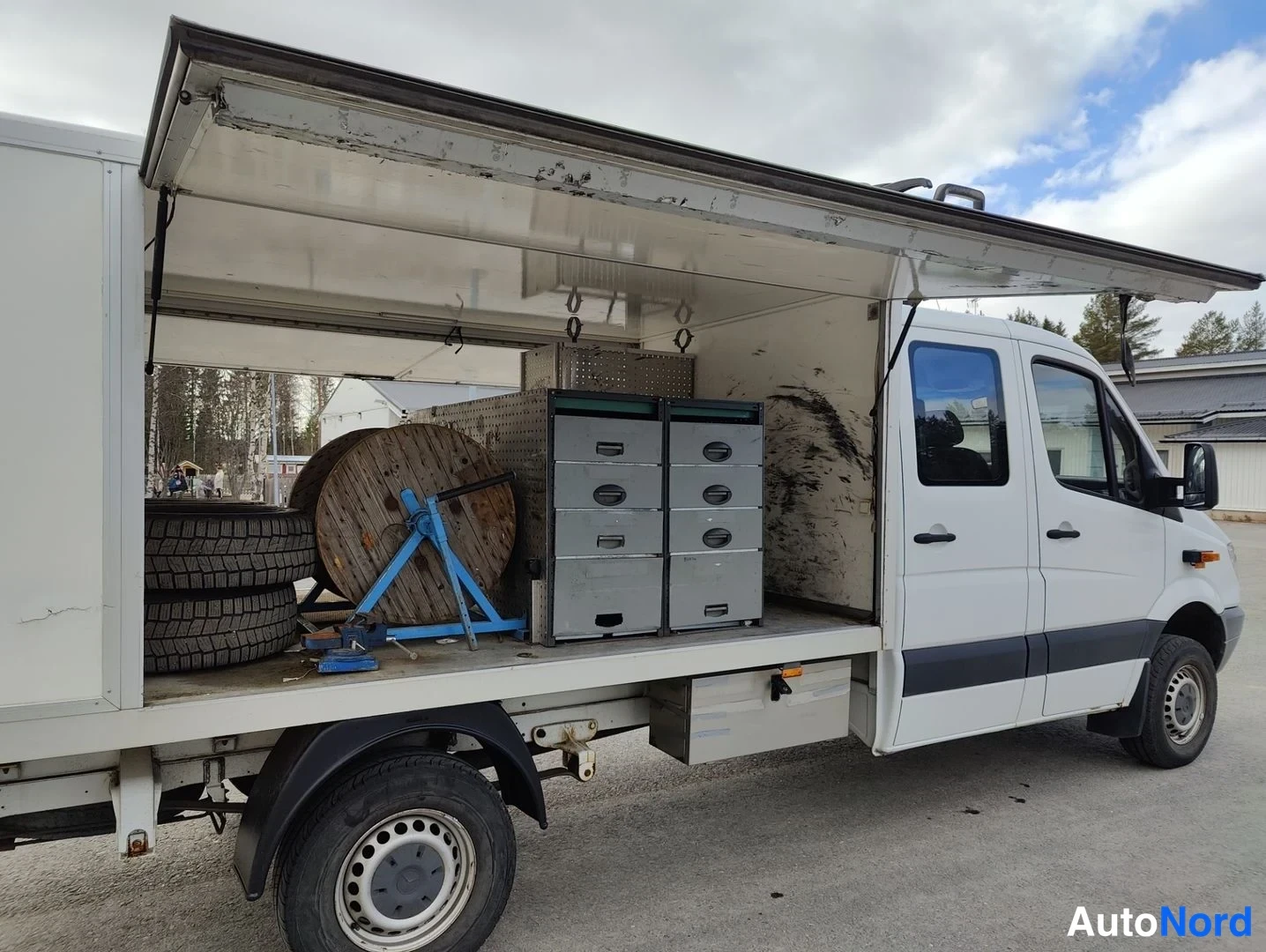 Sivulta: Full left flank view of a white Mercedes-Benz Sprinter panel van used as a work truck. The large cargo body doors are swung open upwards, revealing a flatbed wi