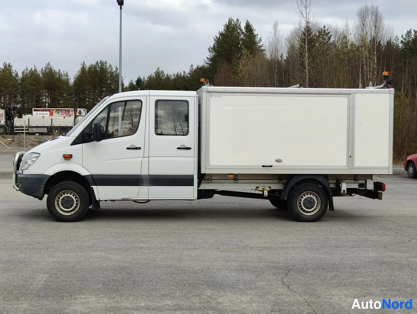 Sivulta: Sivulta nähty valkoinen Mercedes-Benz Sprinter -työauto neliovinen kabiini ja korimalli. Rekisterikilpi näkyy edessä.