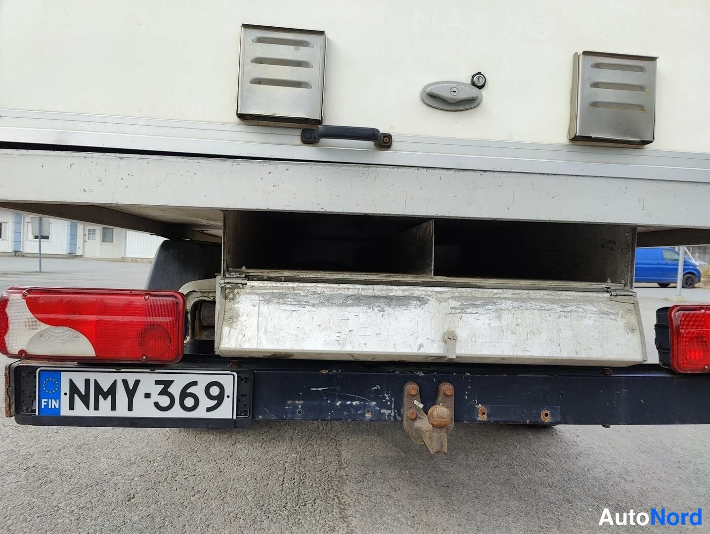 Takaa: Close-up of the rear bumper area with the central under-body storage compartment hinged open, revealing an empty metal space. Two rectangular vents are visible