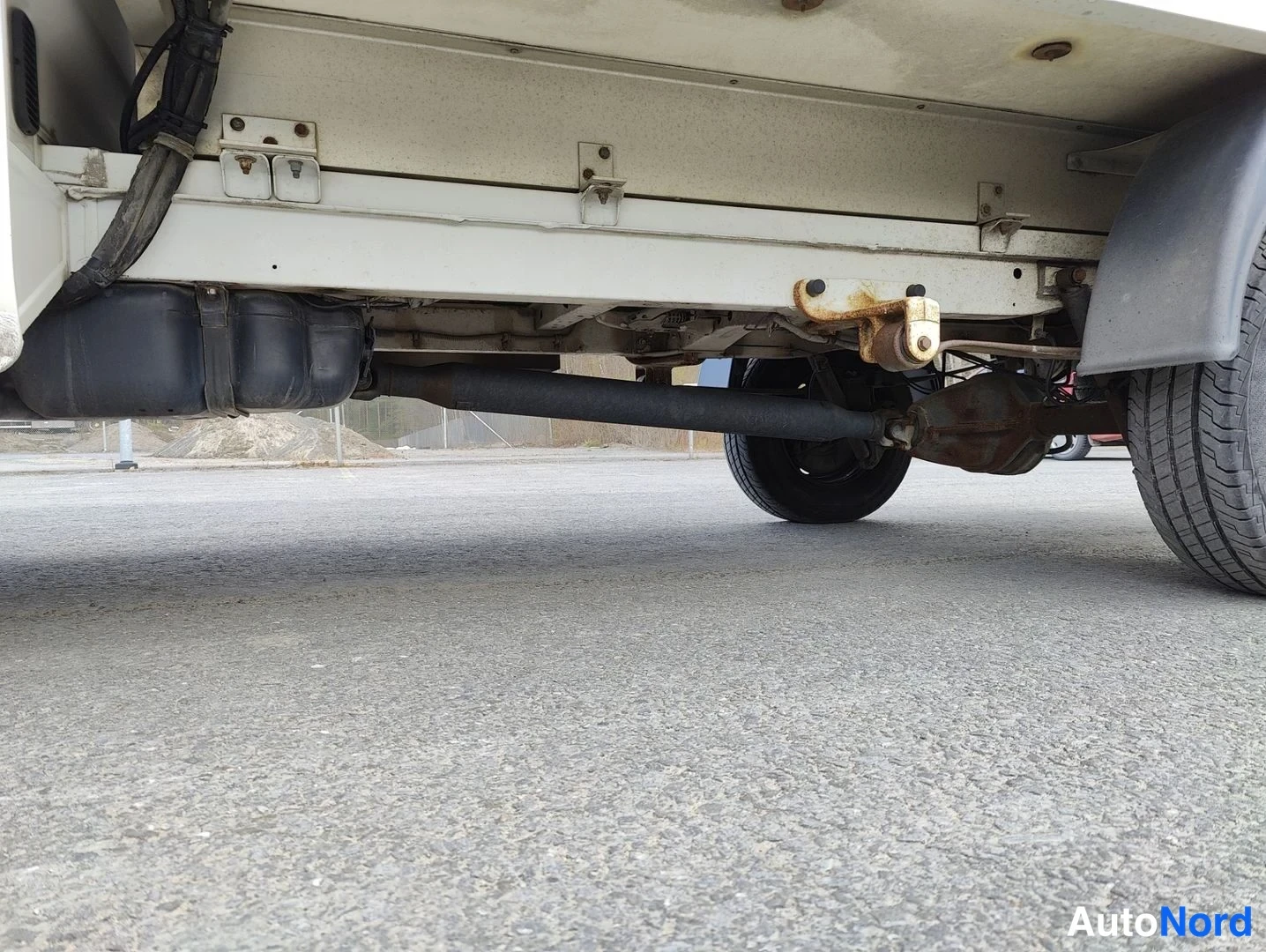 Muu: Low-angle shot underneath the rear of the van showing the solid rear axle, leaf springs, exhaust pipe, and fuel tanks. Rust spots are visible on the chassis fra