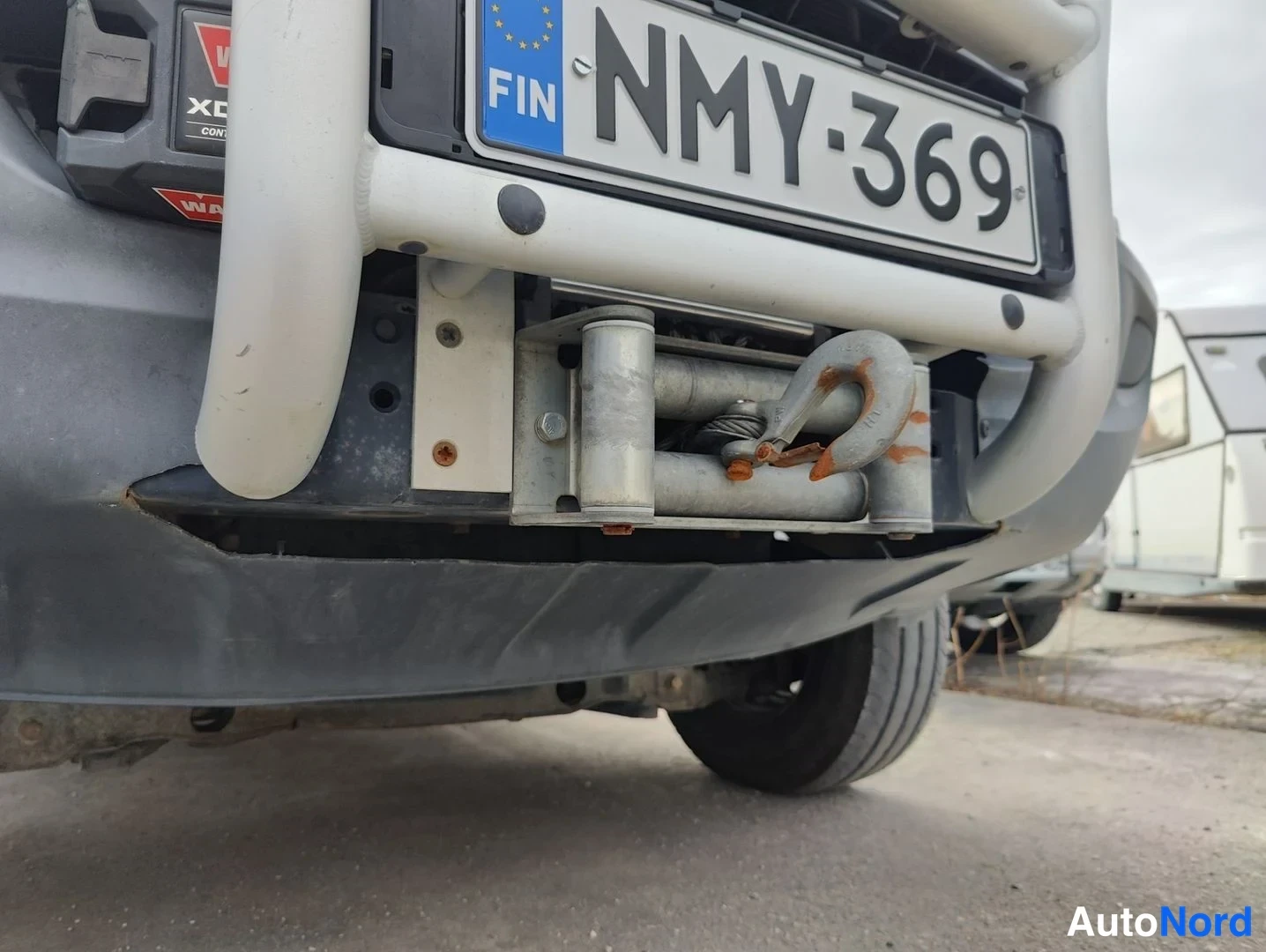Edestä: Close-up front view of a vehicle's lower bumper area showing a Finnish license plate, a white front protective bar with red warning stickers, and an exposed mec
