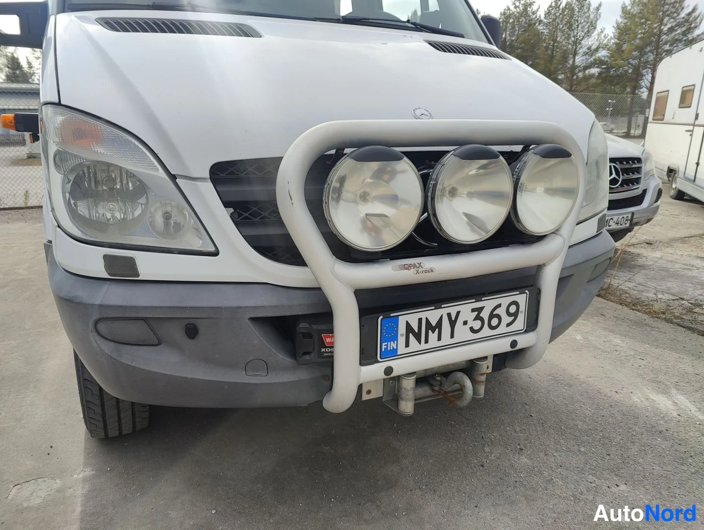 Etuviisto, nokka vasemmalle: Front three-quarter view of a white van with a bull bar fitted over the grille.