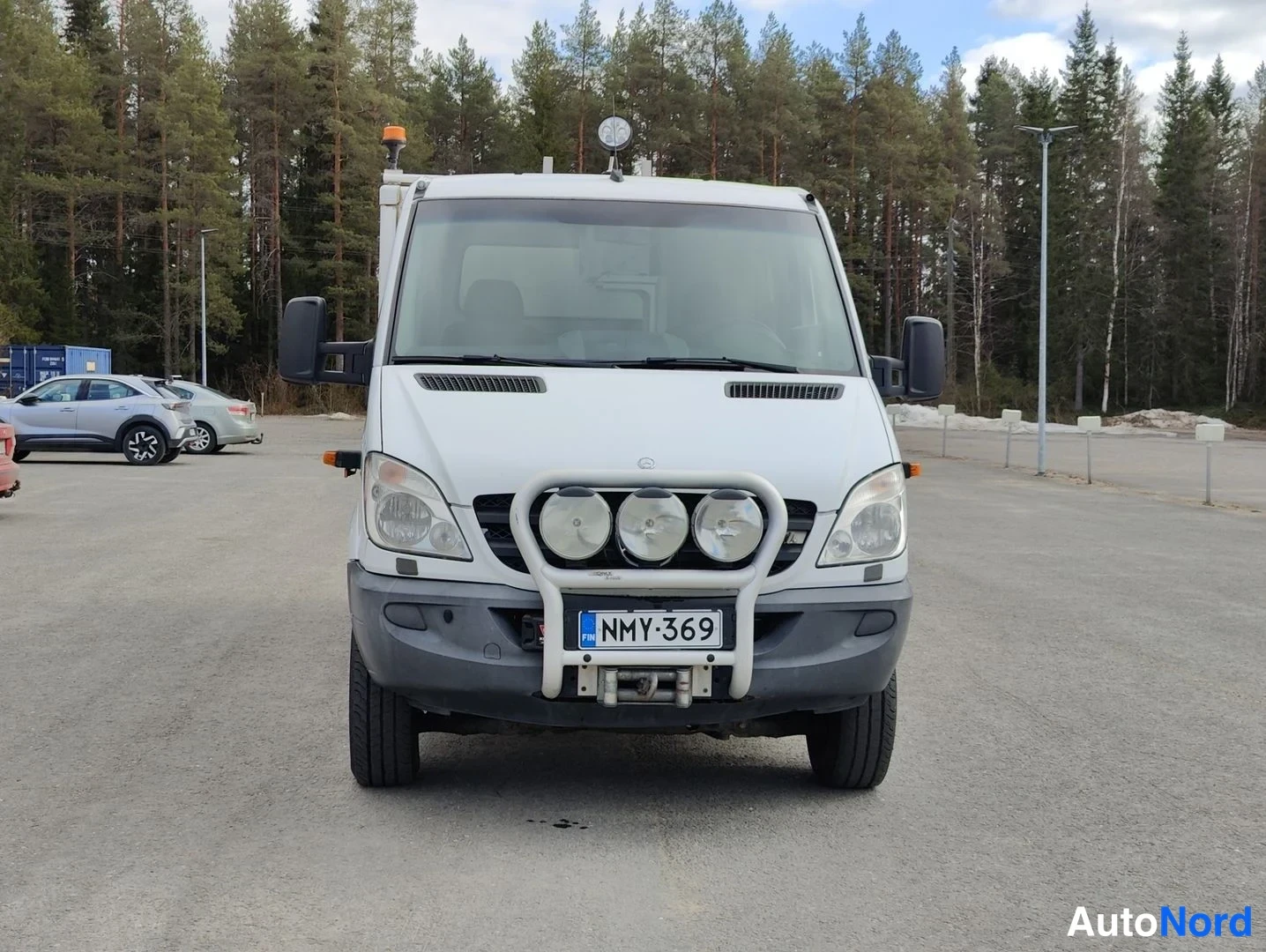 Edestä: Etukuva valkoisesta Mercedes-Benz Sprinterista. Etupuskuriin asennettu suojaputki, jonka kolme suurta pyöreää lisävaloa on kiinnitetty puskurin eteen. Auto on e