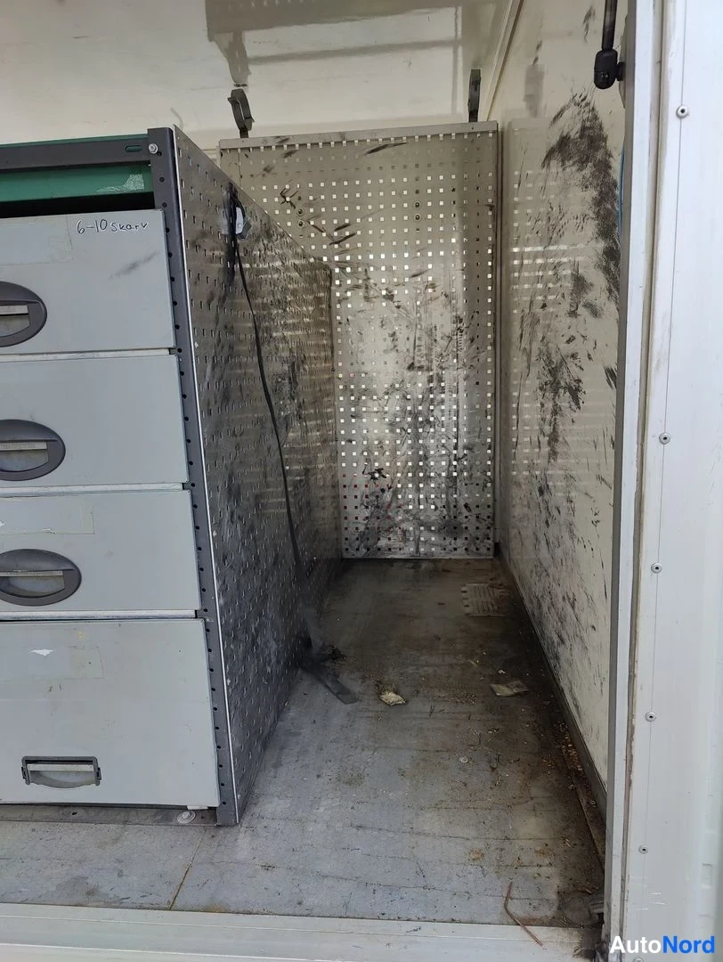Tavaratila: Interior cargo area shot showing a grey metal filing cabinet unit on the left and perforated metal wall panels in the background with black scuff marks. The flo