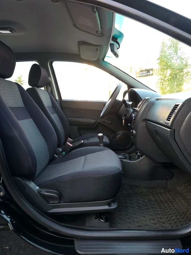 Etuistuimet: Front cabin interior view looking at the driver's side. Shows a three-spoke steering wheel, a manual gear stick, a dashboard air vent, and front seats upholster