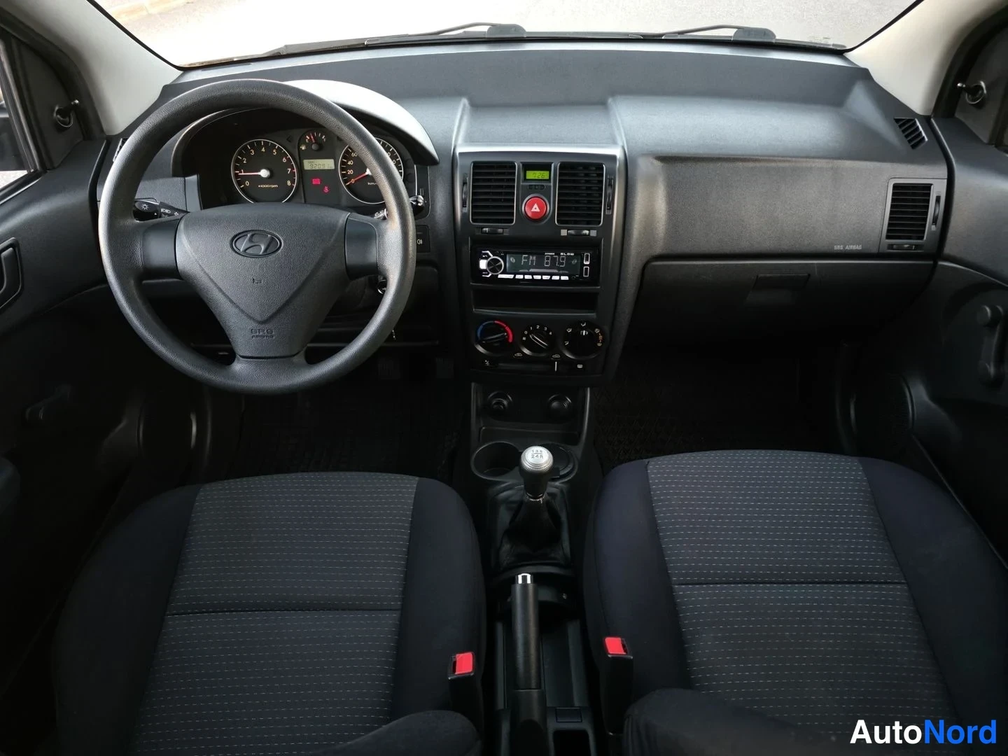 Ohjaamo: Etäkuva auton etusosalta ja ohjaamosta. Mustat kankaan istuimet näkyvät, mukaan lukien selkätukien keskitasot. Ohjauspyörässä on Hyundai-logo ja mittaritaulu nä