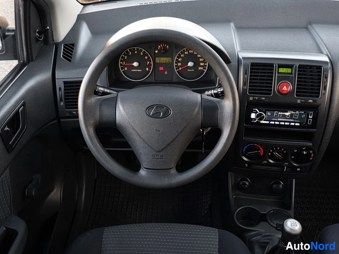 Etuistuimet: Lähikuva ohjauspaneelin ja mittariston alueesta. Mittaritaulussa näkyy nopeusmittari (km/h) ja kierrosluvunmittari. Ohjauspyörän keskellä on Hyundai-logo. Radio