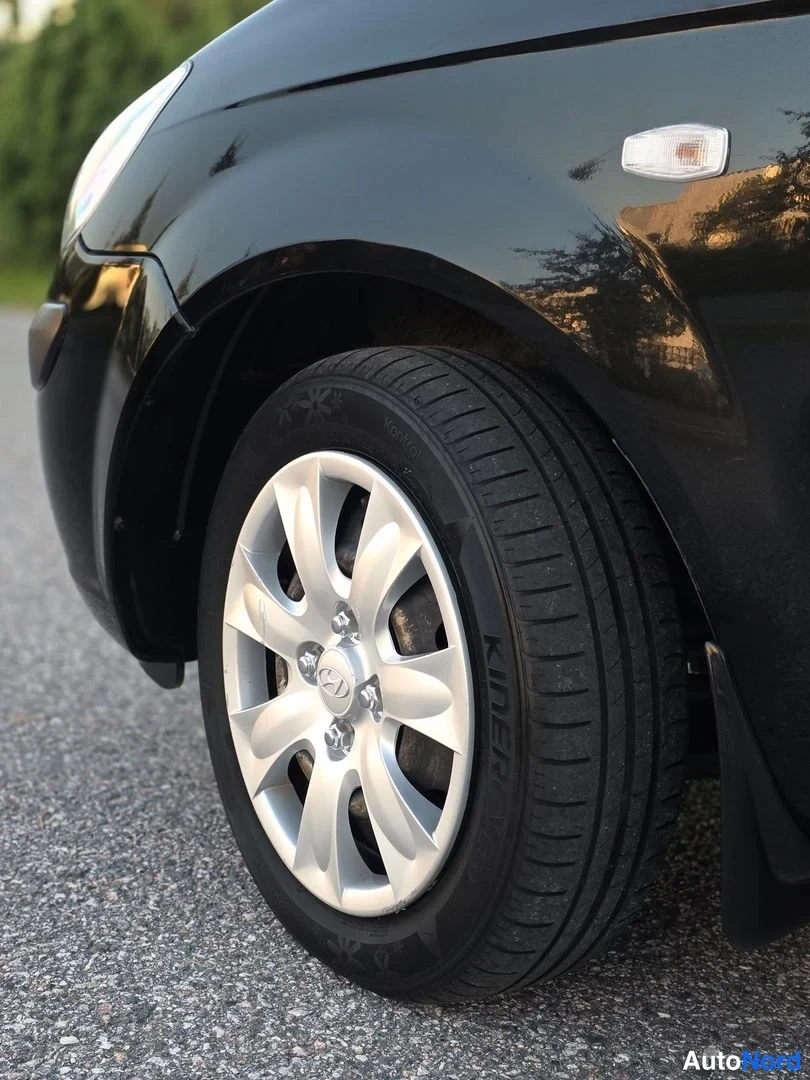 Yksityiskohta: Close-up view of the front left wheel and fender of a black car, showing a silver five-spoke alloy wheel with a central logo, a 'KINEO' branded tyre, and a side