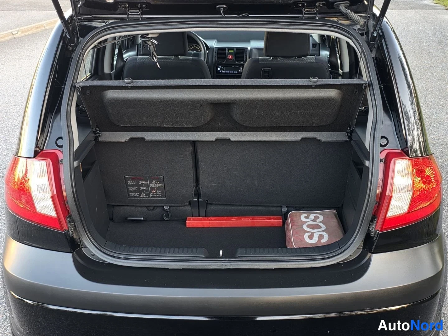 Takaviisto: The open boot of a dark grey hatchback. A parcel shelf divides the space, which contains a red triangular warning sign and an SOS bag. The rear seatbacks are fo