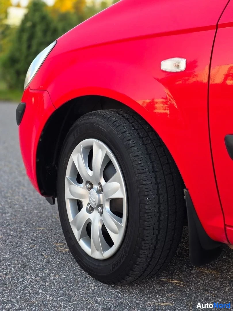 Sivulta: Close-up of a red car's front left side showing the headlight, wheel arch, and a silver alloy wheel with black tyre.