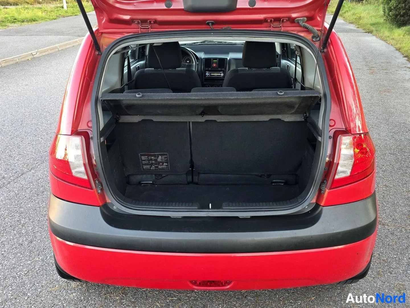 Takaa: Rear view of a red hatchback with the tailgate open, revealing the cargo area with the parcel shelf in place and rear seats folded down.
