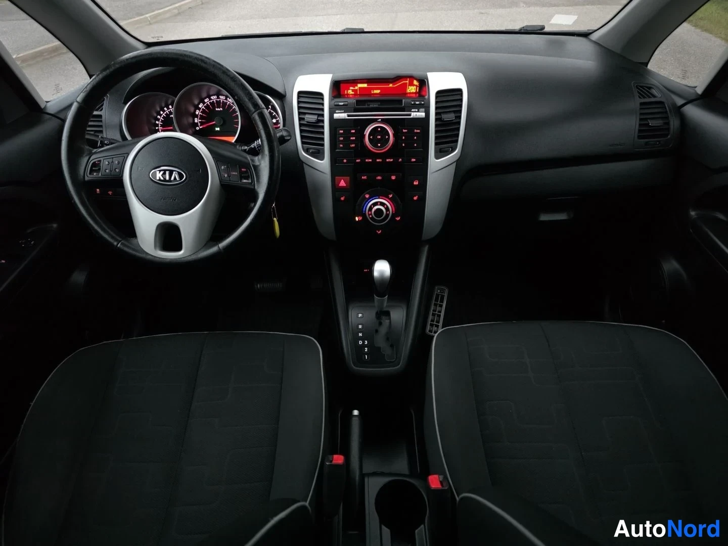 Etuistuimet: Cabin view showing two black fabric front seats, a steering wheel with the Kia logo, an instrument cluster illuminated in red, and a centre console with a silve