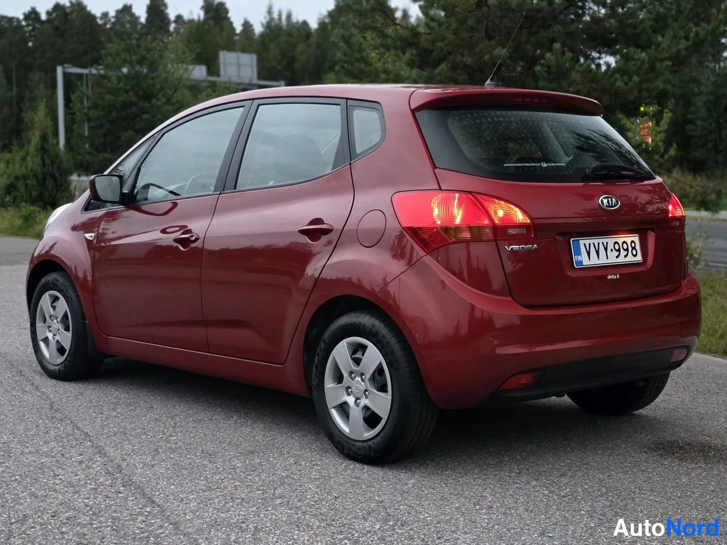 Takaviisto: Tummanpunainen Kia Venga kuvattuna takaa viistosti: takavalo sytytettynä, takaluukussa KIA-merkki ja VENGA-teksti, rekisterikilpi, auto asfaltilla puiden edessä