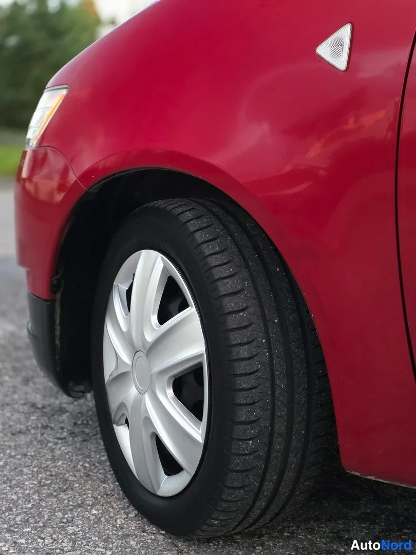 Yksityiskohta: Close-up of a red car's front wheel well showing a silver alloy wheel with five split spokes and black tire on asphalt