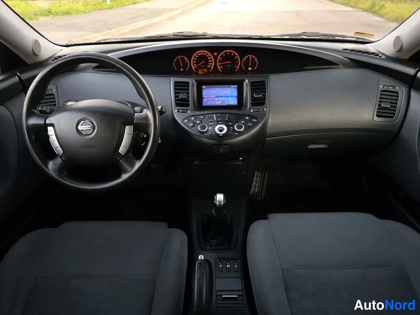 Ohjaamo: Näkymä auton sisälle etupenkkeinä ja keskuspanelina. Ohjauspyörässä Nissan-logo ja monitoimipainikkeet. Mittaritaulu kolmella mittarilla oranssilla valolla. Kes