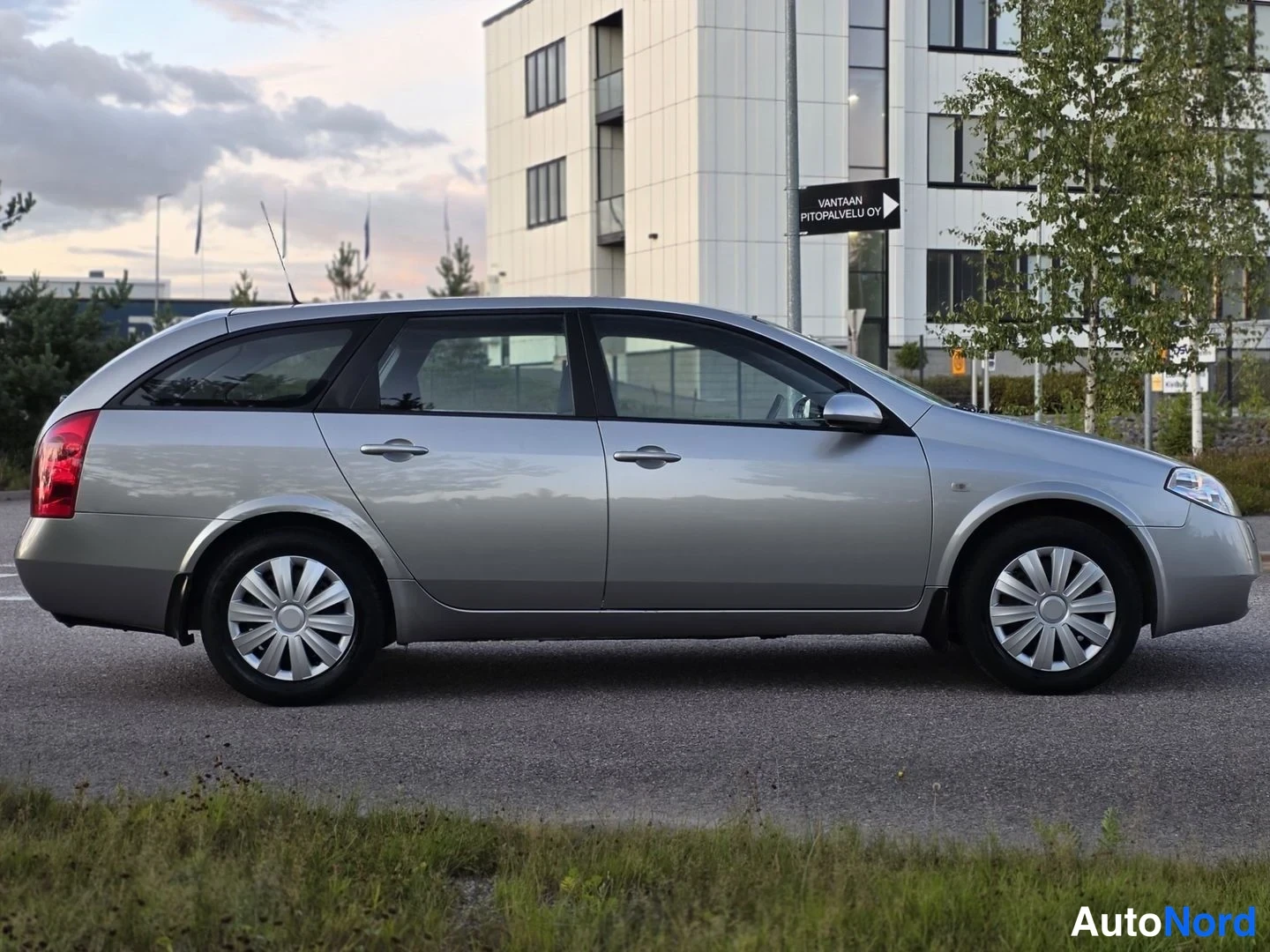 Sivulta: Silver Nissan station wagon photographed in strict side profile from the left. Shot on asphalt with grass and a building in background.