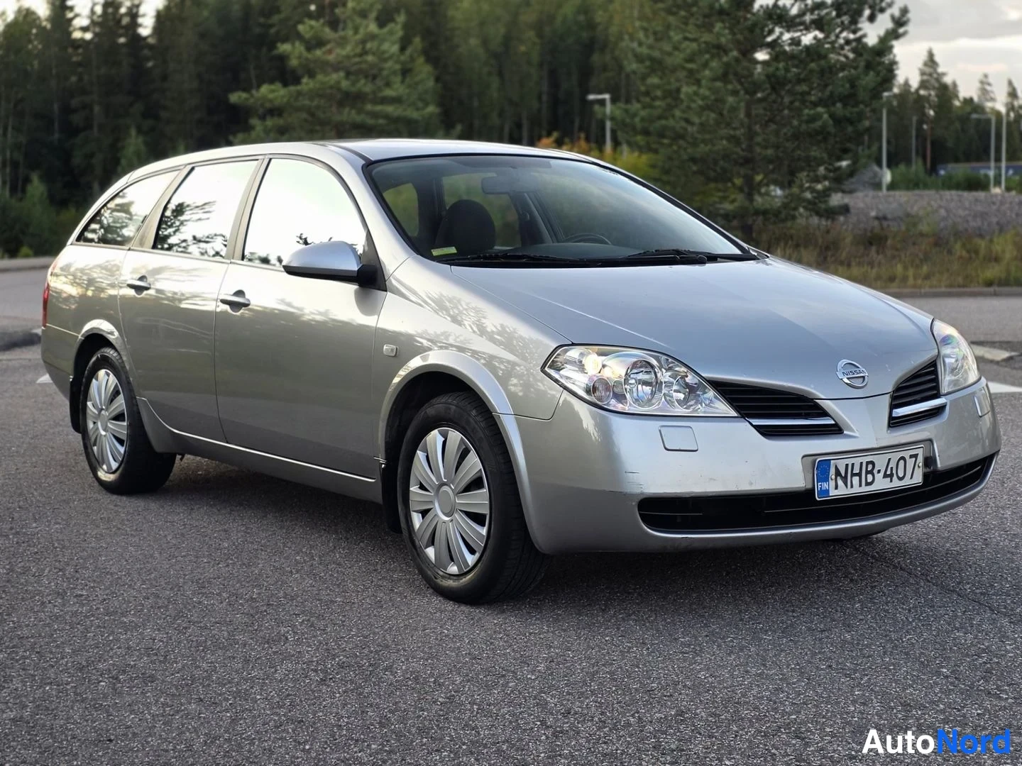 Etuviisto, nokka vasemmalle: Front three-quarter view of silver Nissan station wagon showing front face and driver side flank. Front right headlight on.