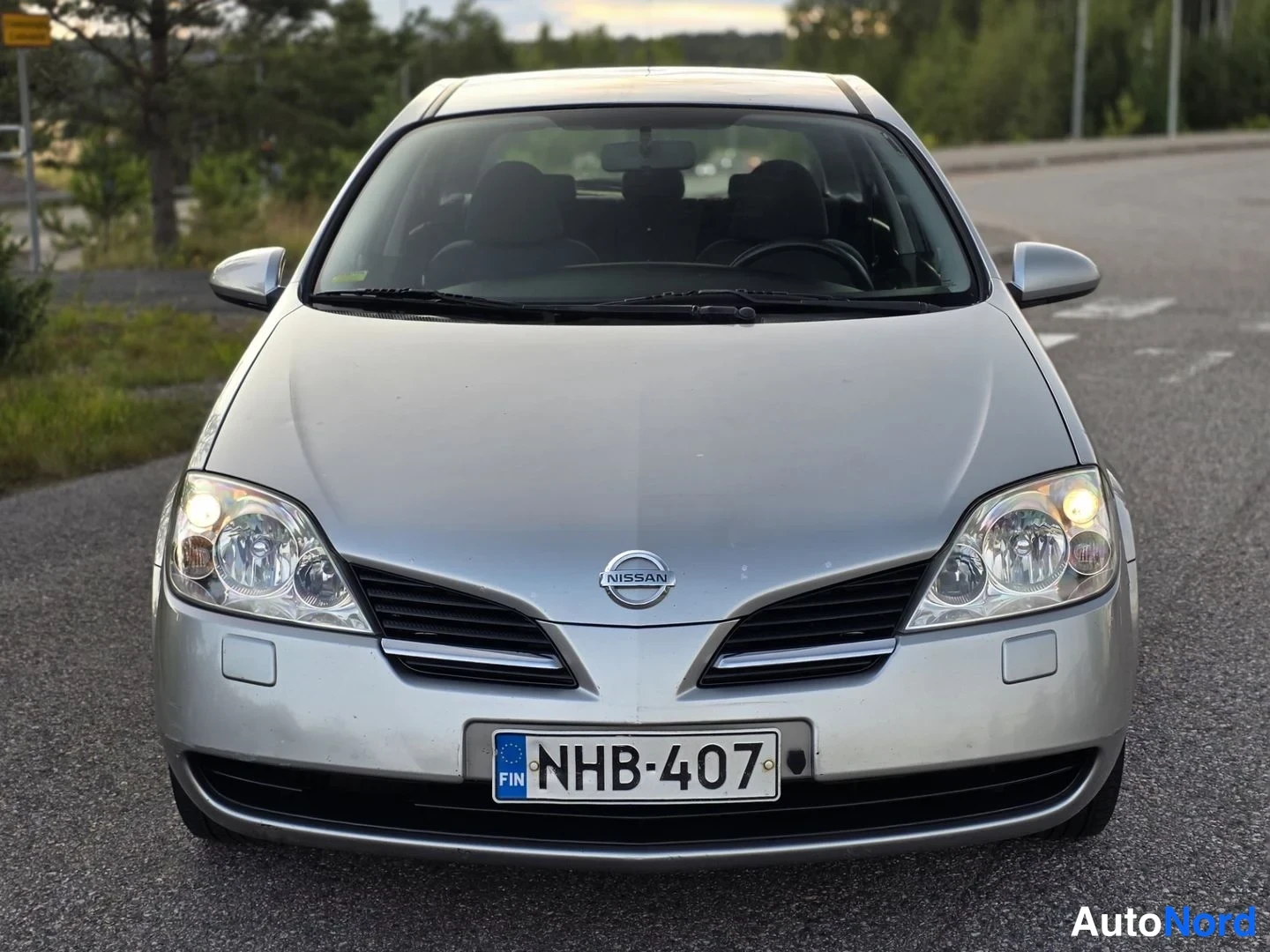 Edestä: Direct frontal shot of silver Nissan displaying grille, Nissan emblem, headlights, fog lights and license plate.