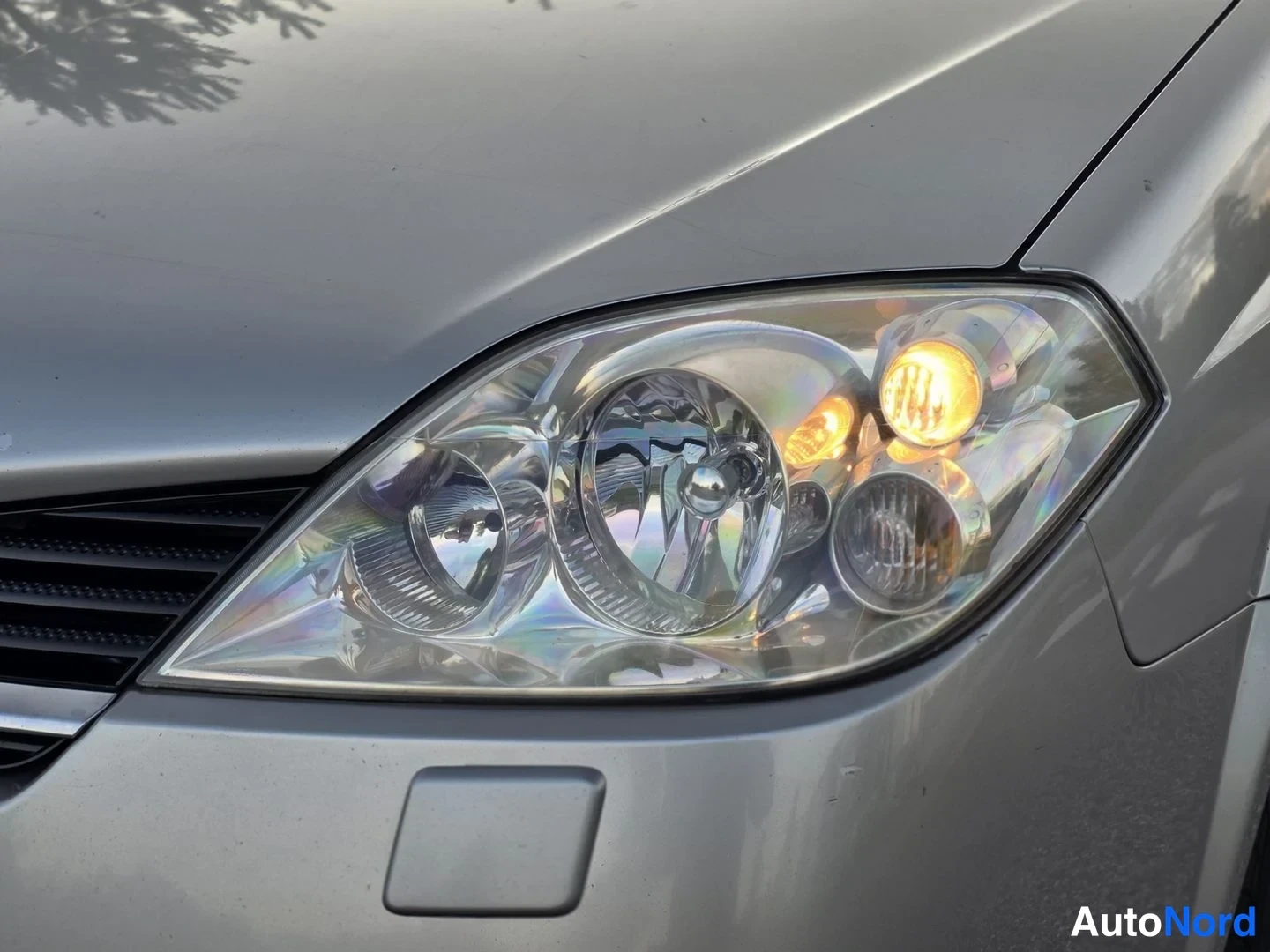 Yksityiskohta: Close up of front right headlamp assembly with amber turn indicator lit. Adjacent square grey area is parking sensor housing. Visible portion includes black gri
