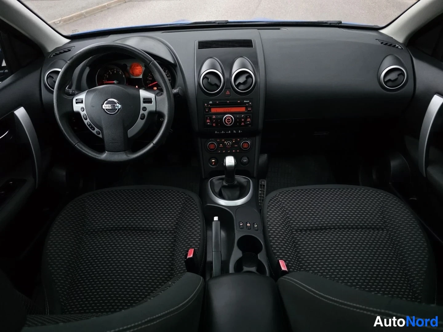 Etuistuimet: Etualtakulma auton sisätilaan. Mustat kankaan istuimet etualalla. Ohjauspyörä Nissan-logolla ja vaihteenvalitsin keskipaneelissa.