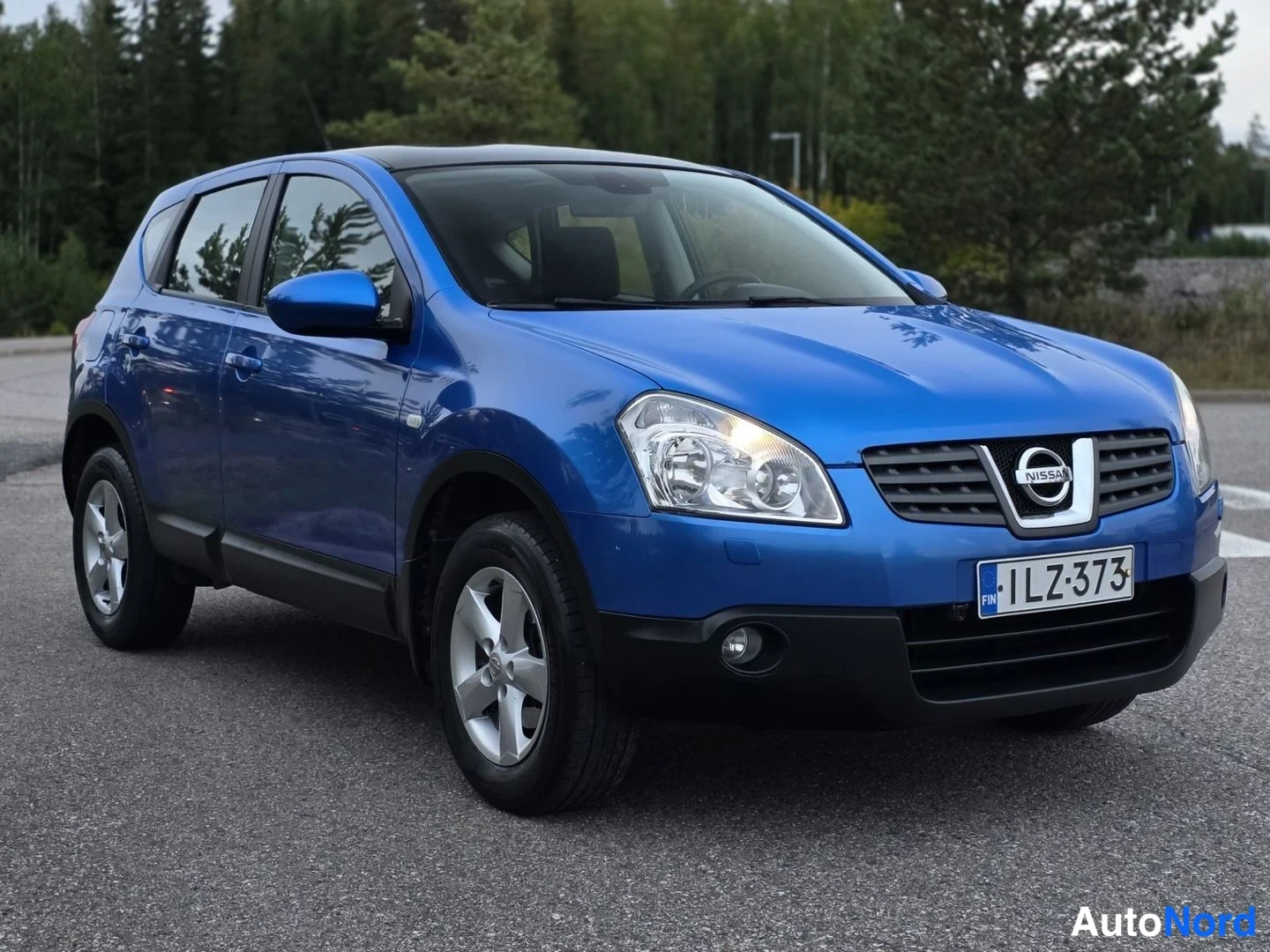 Etuviisto, nokka oikealle: Edestä ja oikealta kuvattu sininen Nissan Qashqai, etusäleikkö ja kylki näkyvissä, rekisteritunnus.