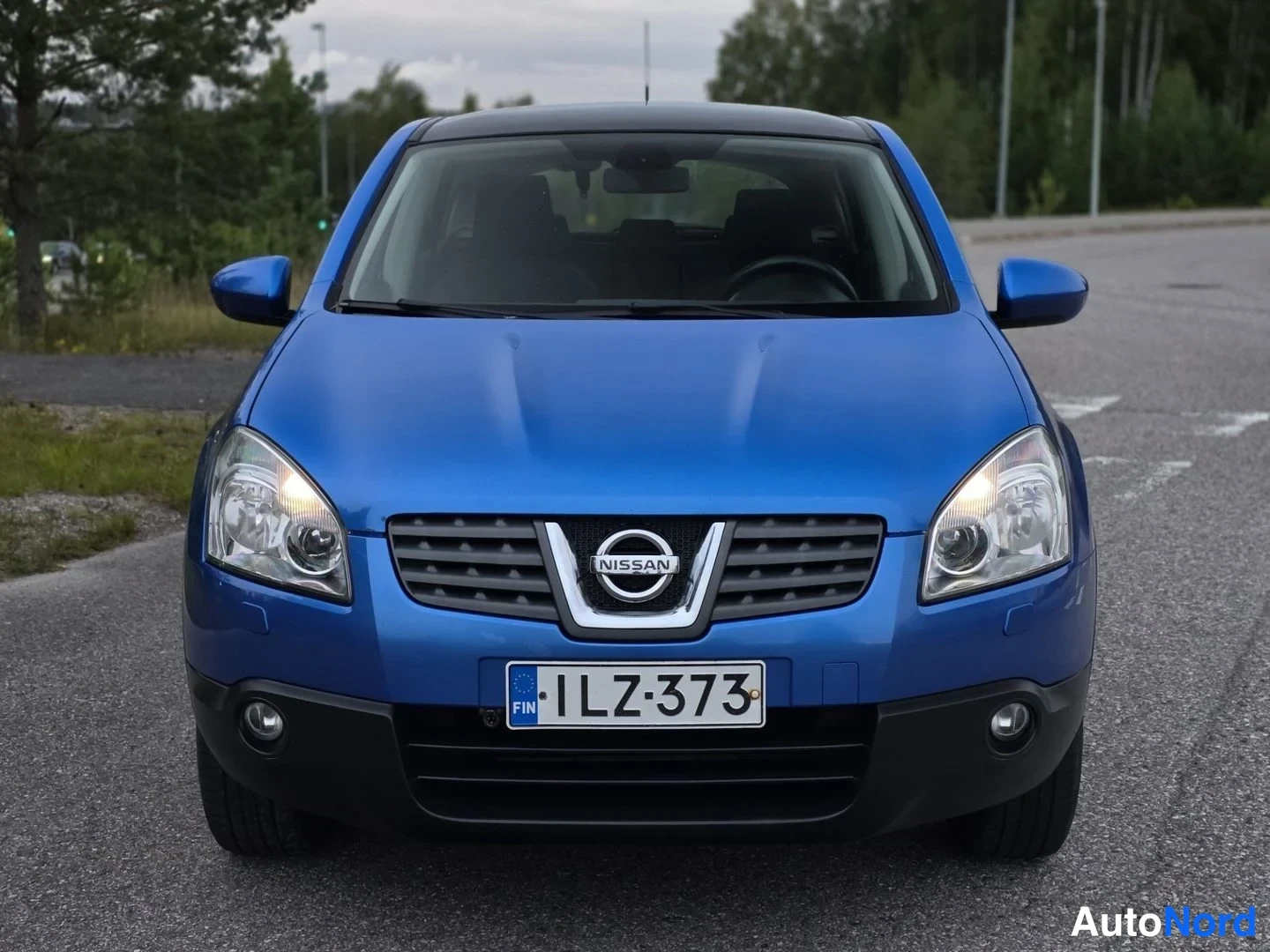 Edestä: Suoraan edestä kuvattu sininen Nissan Qashqai, ajovalot päällä, rekisteritunnus.