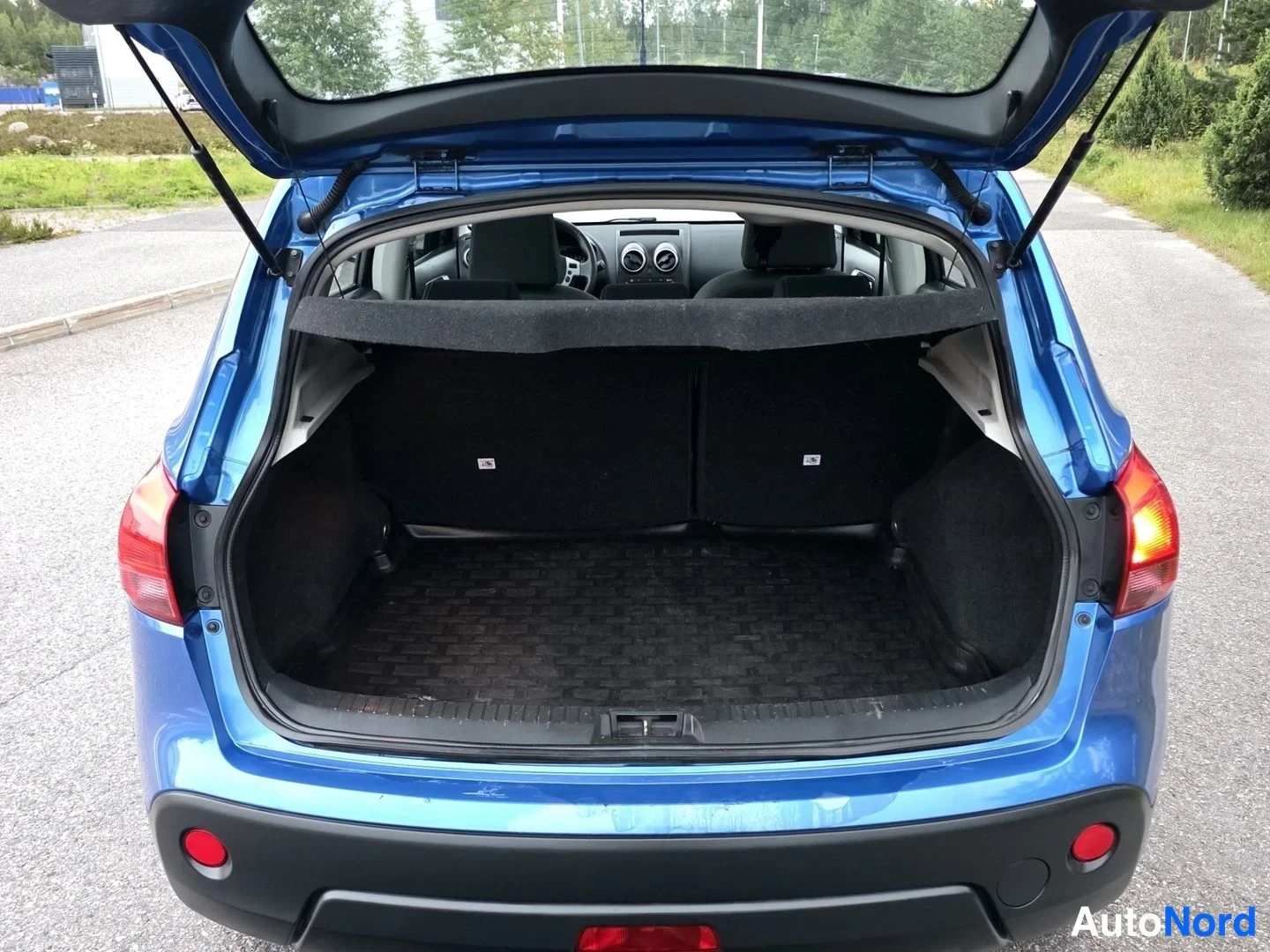 Takaviisto: Rear view of a blue car with its tailgate open, revealing an empty cargo area with a black cover and parcel shelf. Through the opening, front seats, steering wh