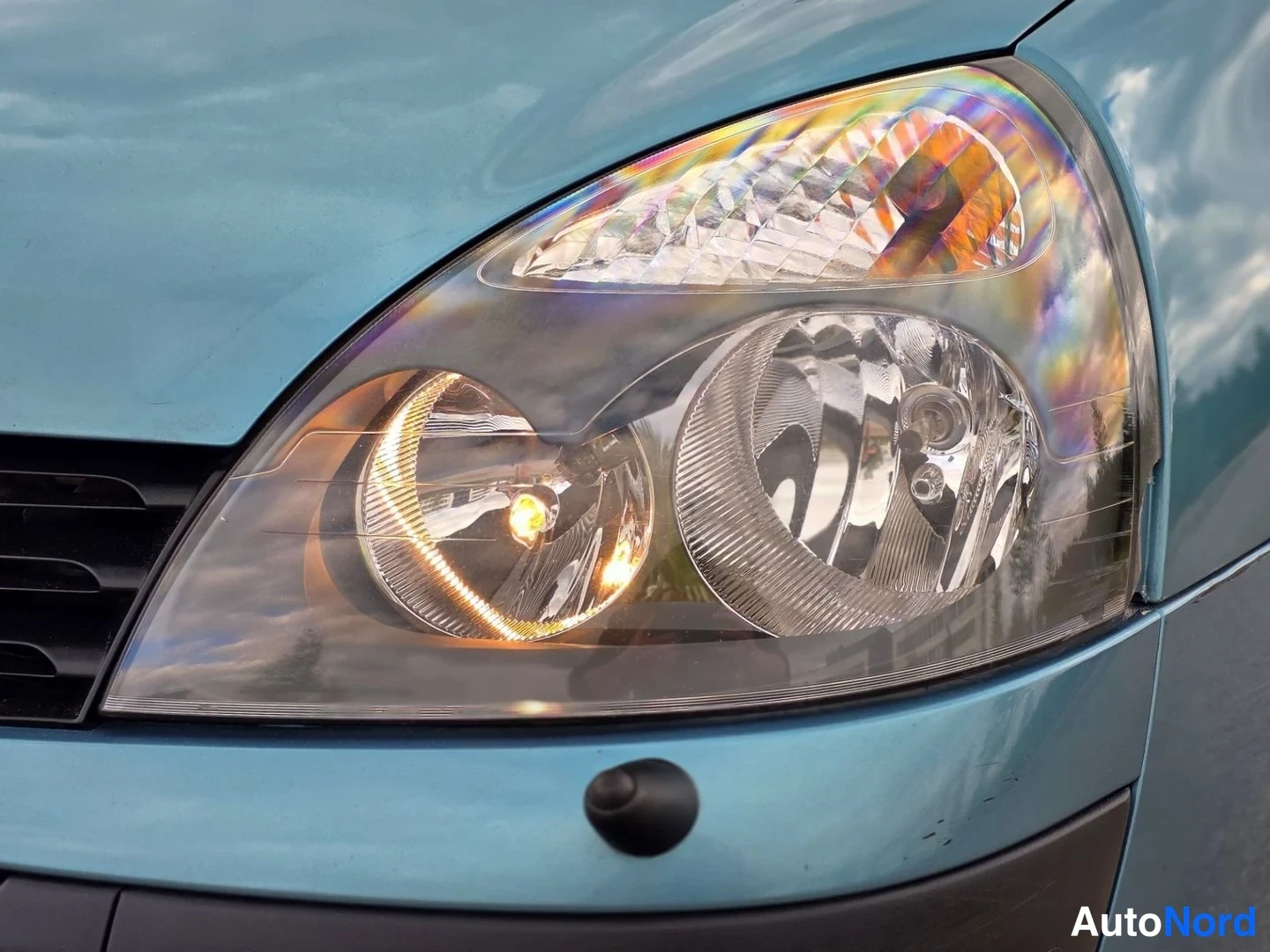Yksityiskohta: Close-up of the driver's side headlamp assembly showing main beam, fog lamp and amber indicator with rainbow refraction pattern in lens.
